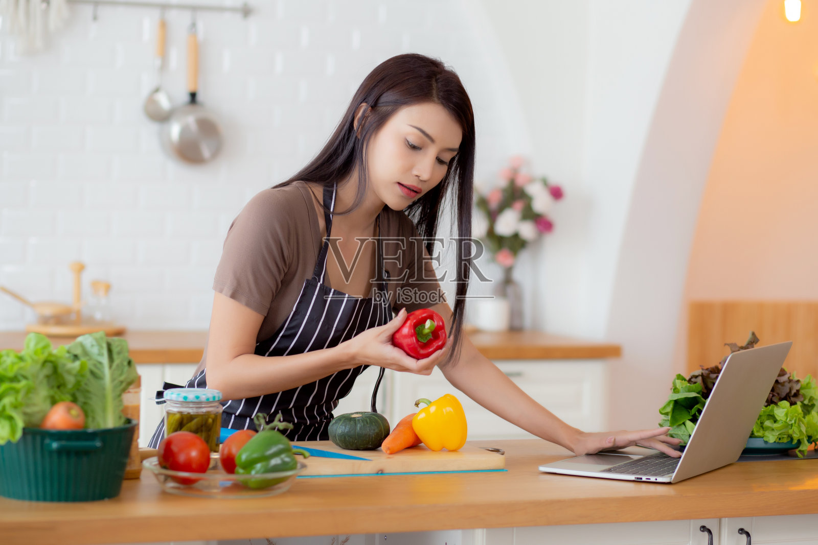 年轻的亚洲女性在家里的厨房里一边做沙拉蔬菜，一边拿着甜椒看笔记本电脑，快乐的女性为健康准备食物的食材，看着笔记本看食谱。照片摄影图片