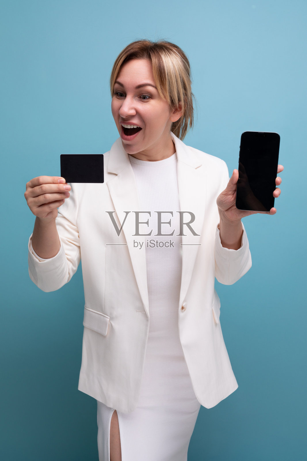 自信的年轻欧洲女子穿着白色夹克和裙子，为她的银行卡和智能手机做广告照片摄影图片