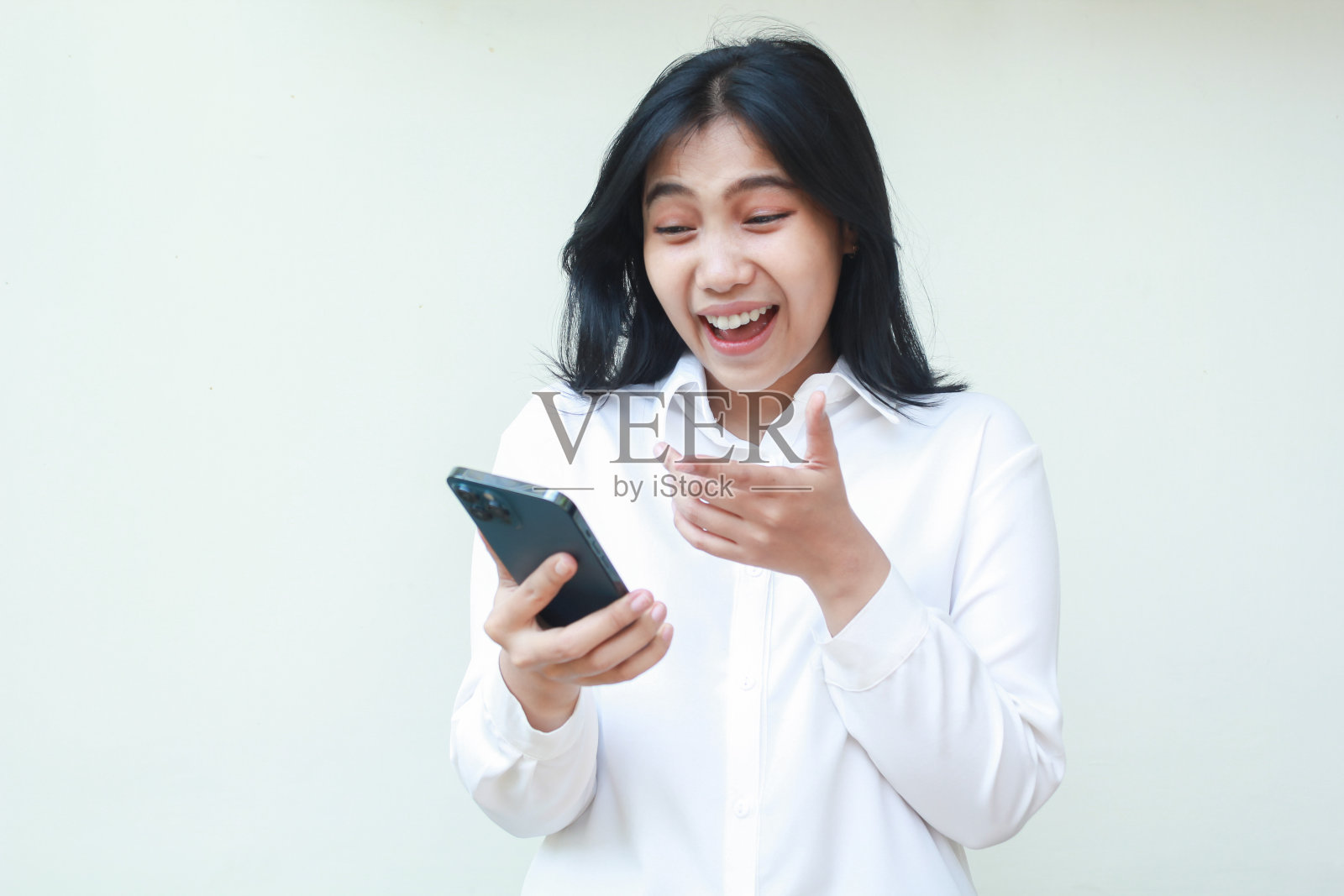 无忧无虑的亚洲年轻商业女经理的形象，穿着白色正装衬衫，举起手掌，孤立地站着，在智能手机上笑着照片摄影图片