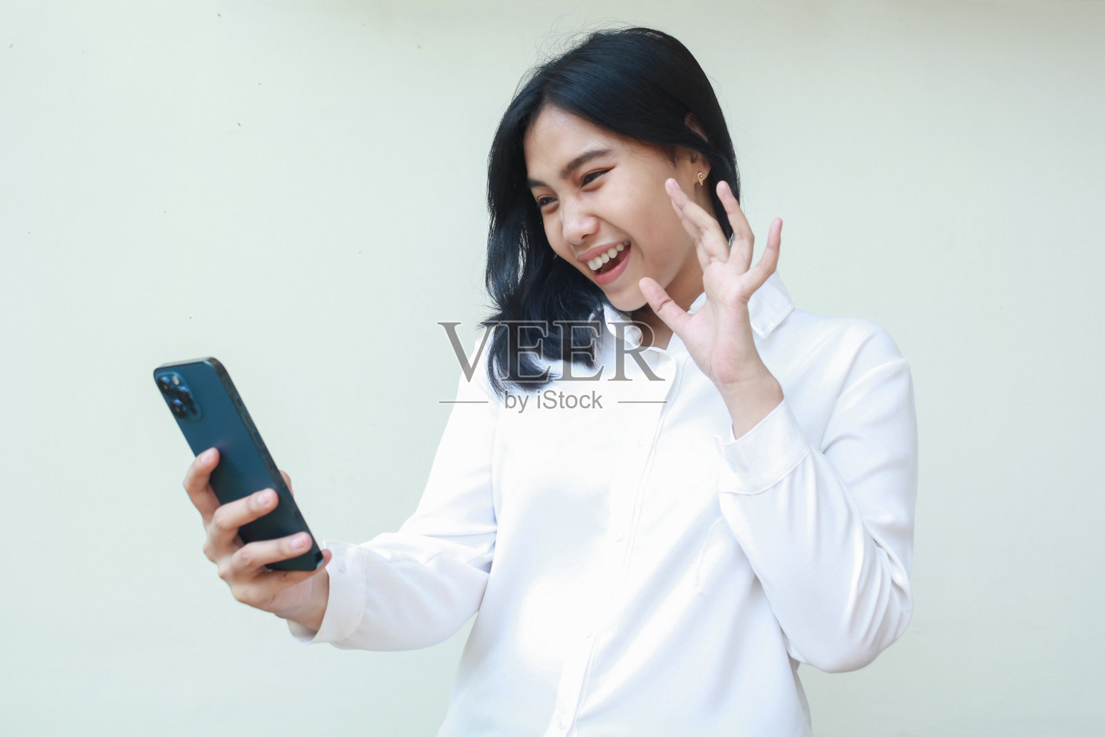 照片中，年轻的亚洲女企业家拿着智能手机，微笑着举手打招呼，穿着白色西装衬衫，秘书通过视频电话表示欢迎，孤立地站着照片摄影图片