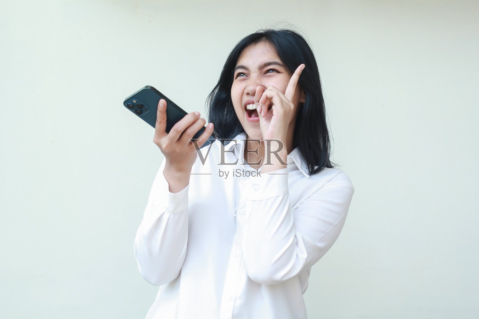 欣喜若狂、无忧无虑的亚洲年轻女售货员讲着手机，双手紧握，望向别处笑着，手指指向拷贝空间，穿着白色正装衬衫，孤立地站着照片摄影图片