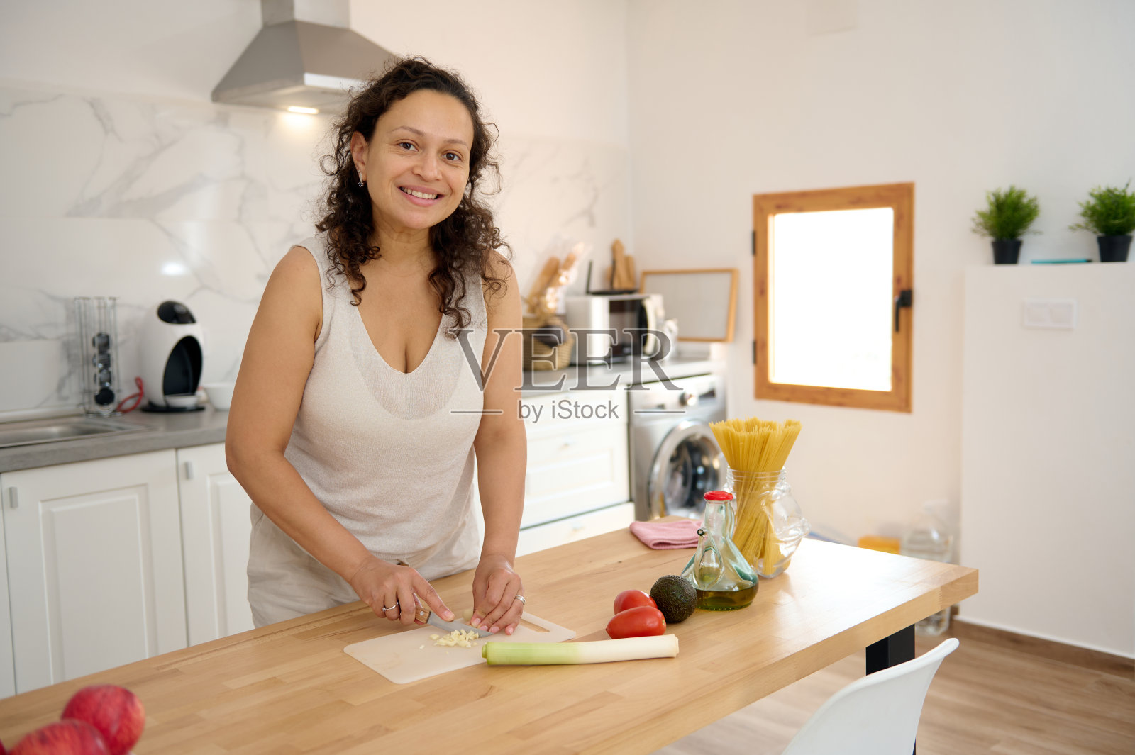 在极简主义的家庭厨房里，美丽的家庭主妇微笑着看着镜头，在切菜板上切韭菜和大蒜照片摄影图片