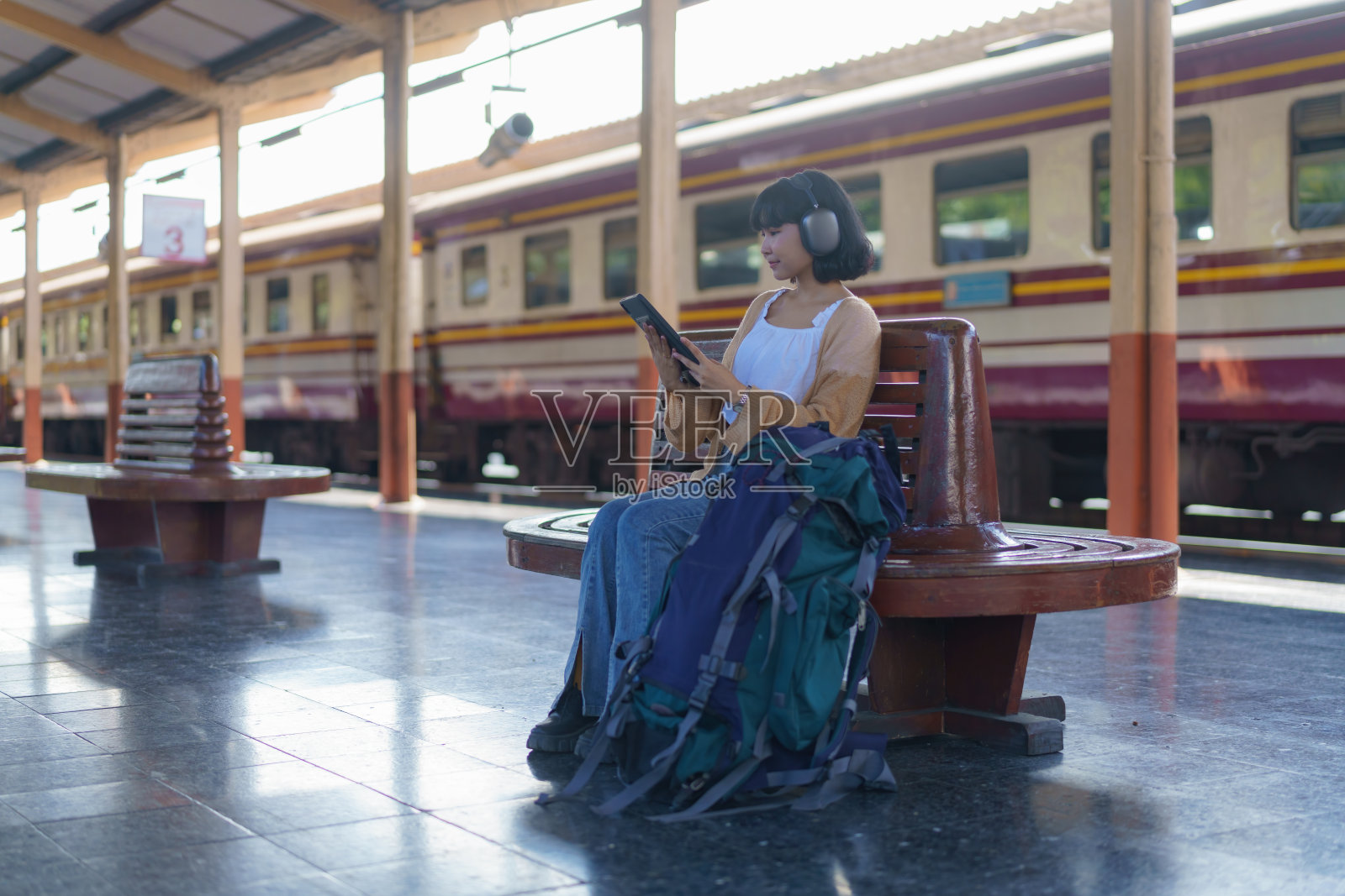 一位亚洲女游客，坐在火车站的长椅上，戴着耳机，听着平板电脑上的音乐，等待火车的到来。照片摄影图片