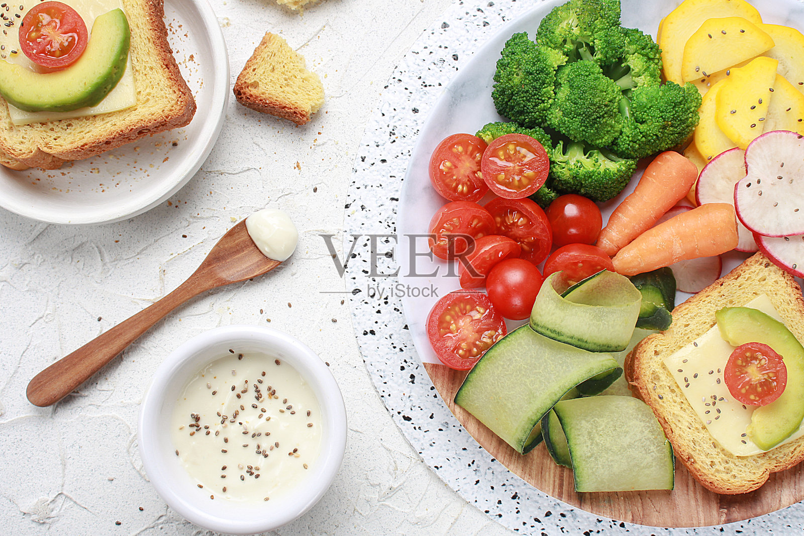 简单的午餐或晚餐，有樱桃番茄、黄瓜、西葫芦、胡萝卜、西兰花、萝卜和烤面包、鳄梨和奶酪，放在白色的桌子上。照片摄影图片