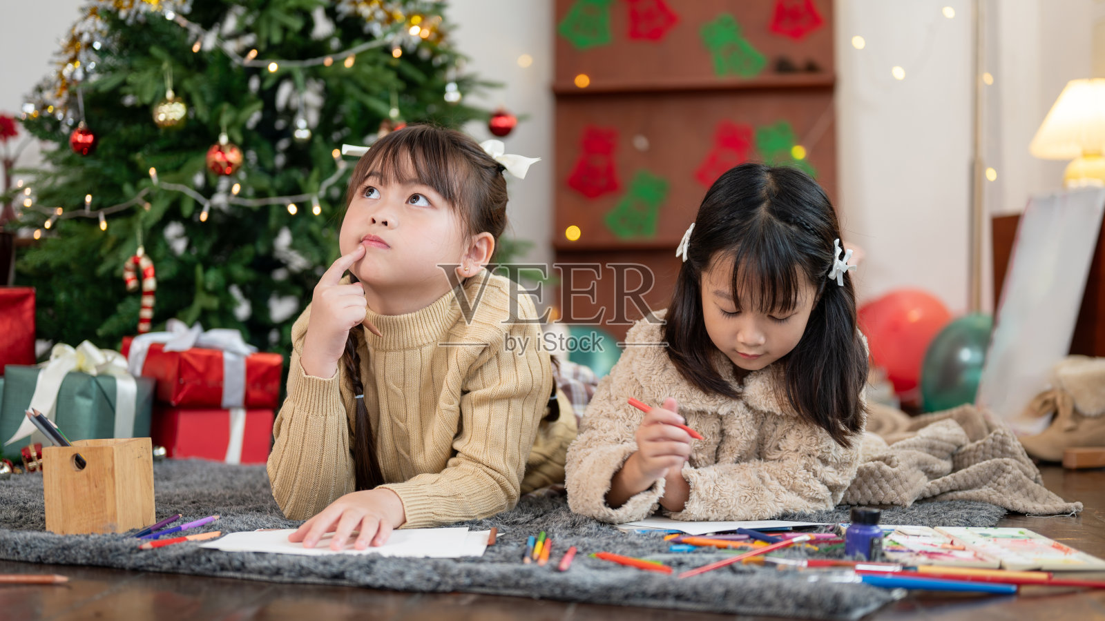 两个可爱的年轻亚洲女孩躺在圣诞树旁边的地板上一起涂色。照片摄影图片