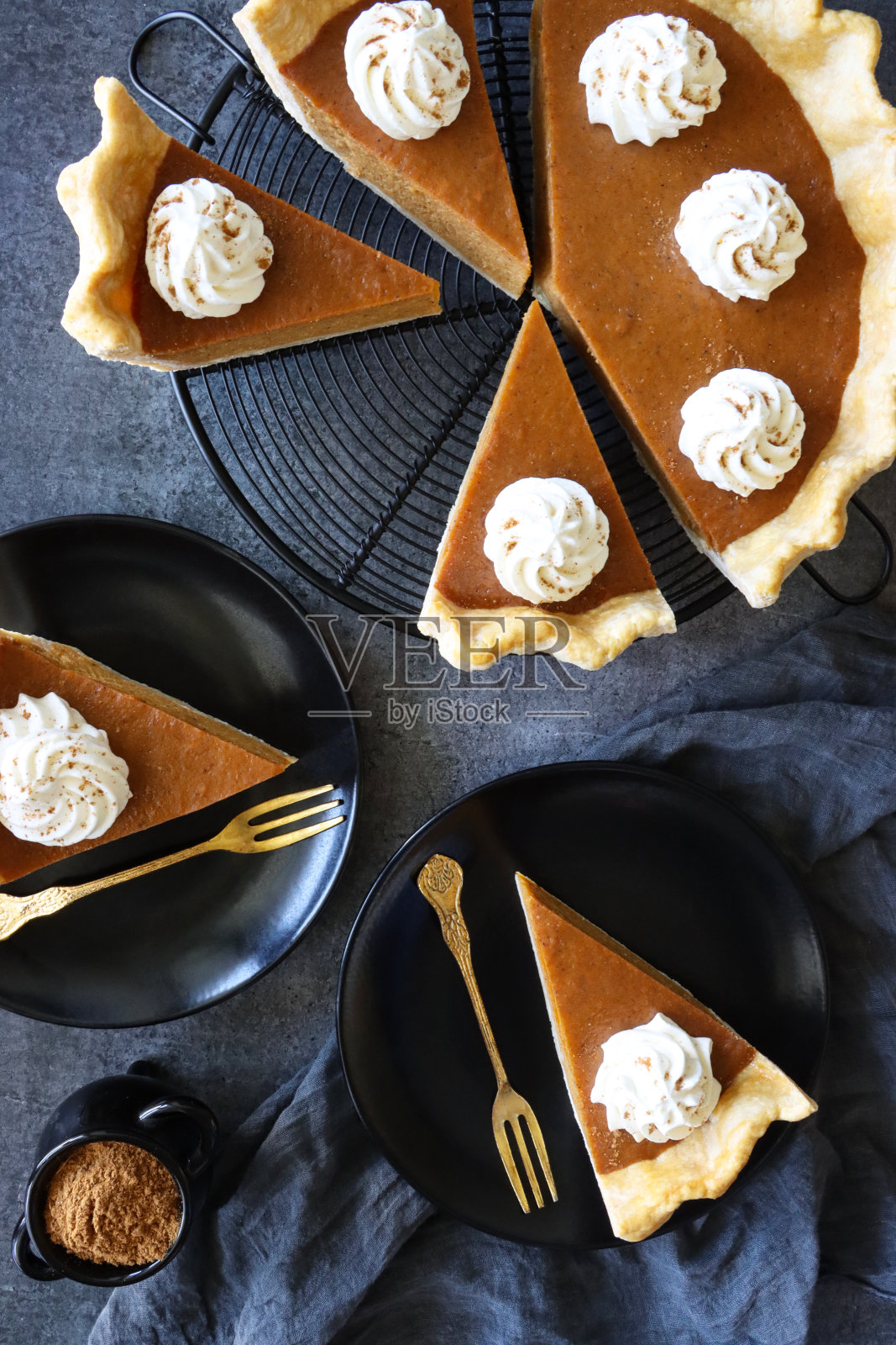 近景:黑色盘子里放着新鲜出炉的万圣节南瓜派，上面撒着漩涡状的鲜奶油，冷却架上放着带凹槽的馅饼皮，里面装满了南瓜泥，一锅混合香料粉，金色的叉子，灰黑色的细布，高架视图照片摄影图片