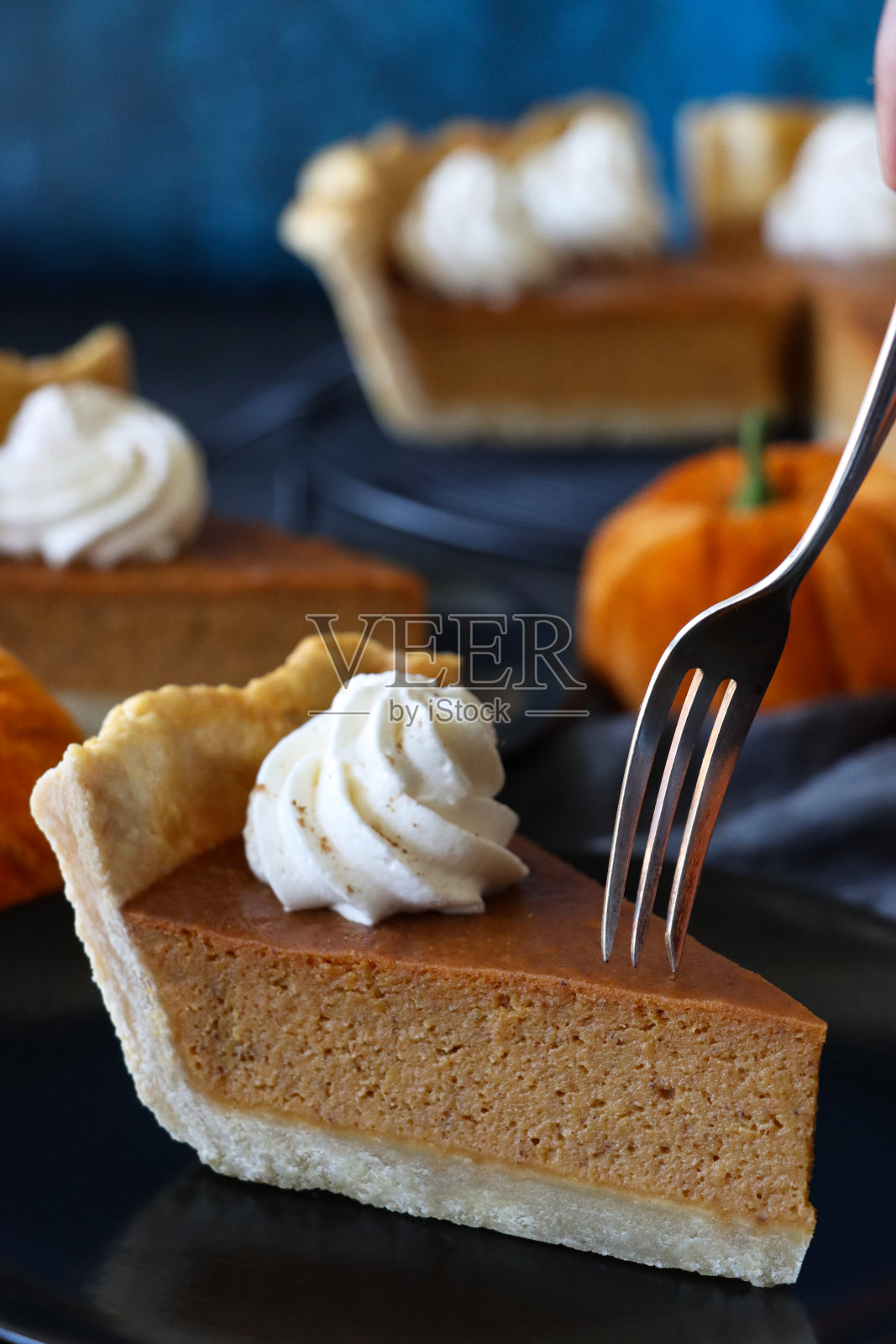 黑色盘子上的叉子切成一片新鲜出炉的万圣节南瓜派，上面有搅打过的奶油，毛绒南瓜，在冷却架上有凹槽的馅饼皮，里面装满了南瓜泥，蓝色背景斑驳照片摄影图片