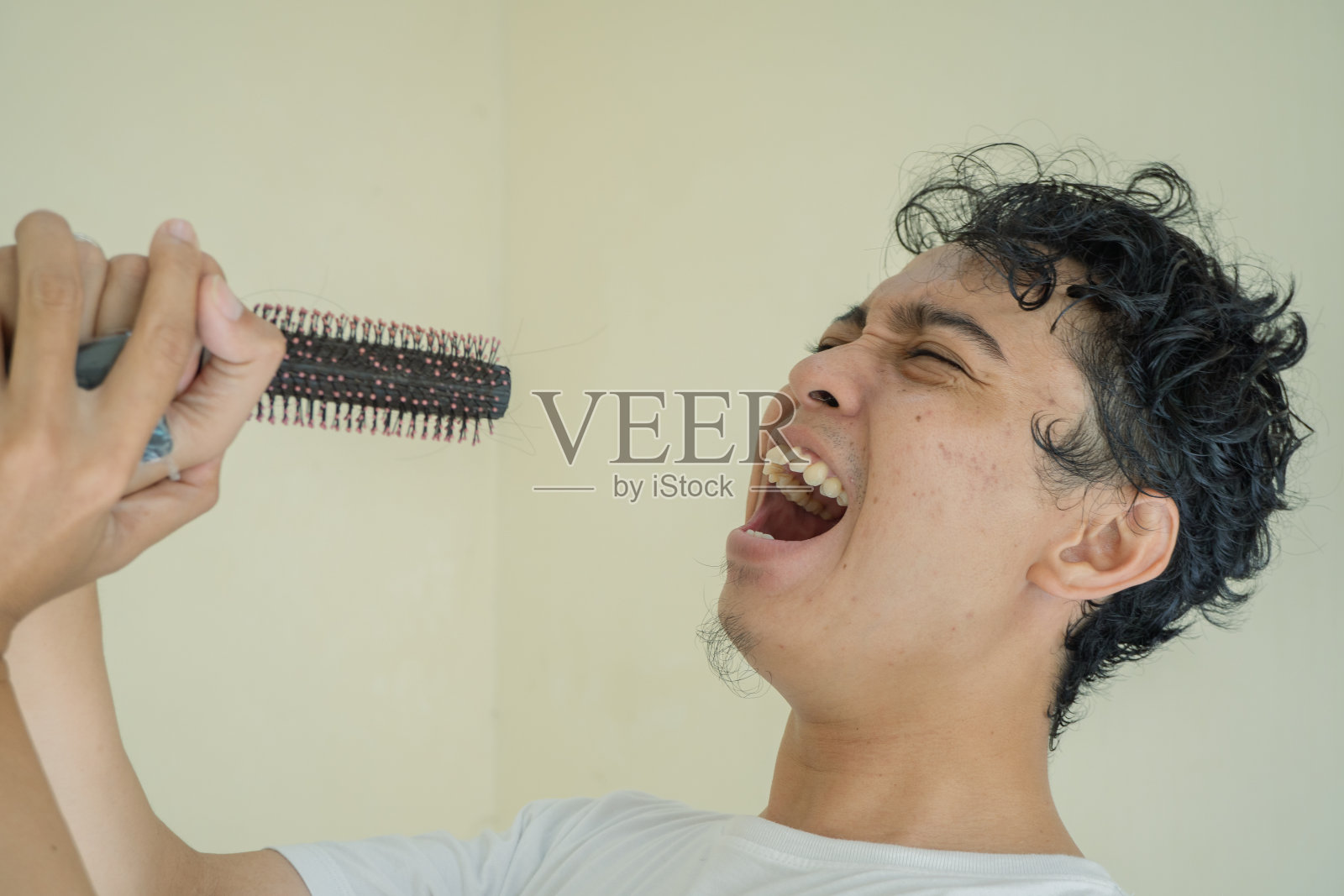 年轻卷曲的男子穿着白色t恤唱歌时用梳子做手势。照片摄影图片