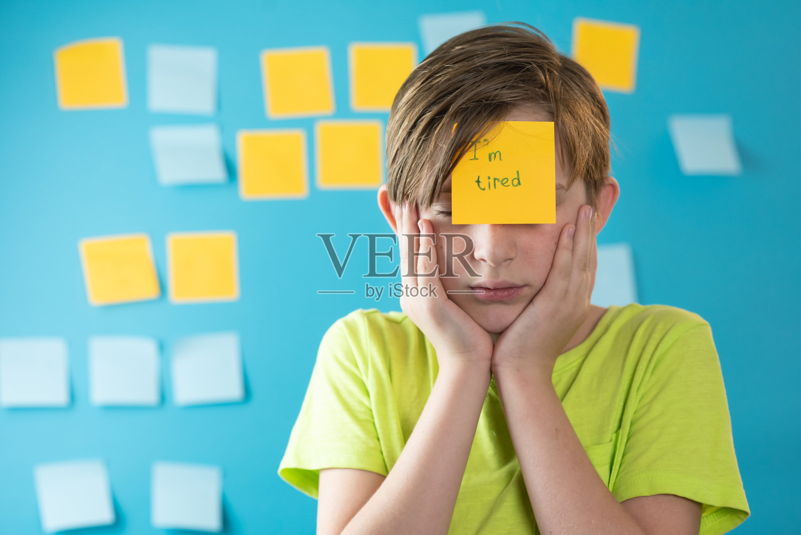 一个疲惫的11岁男孩站在贴有贴纸的蓝色墙壁前。额头上贴着“我累了”贴纸的严肃儿童照片摄影图片