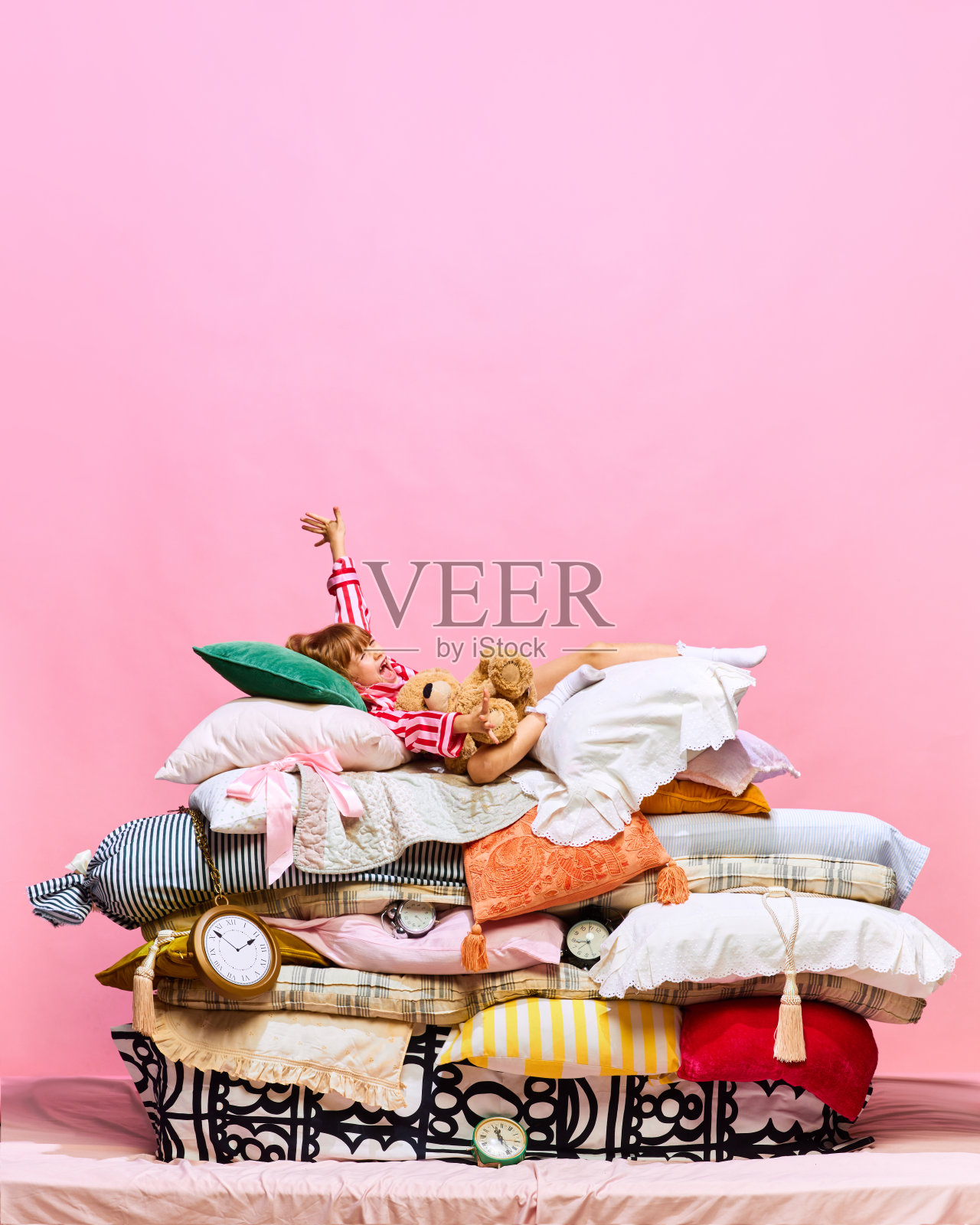 早上好。可爱的小公主和可爱的泰迪熊躺在大羽毛床上的肖像，粉红色的工作室背景上有枕头。照片摄影图片