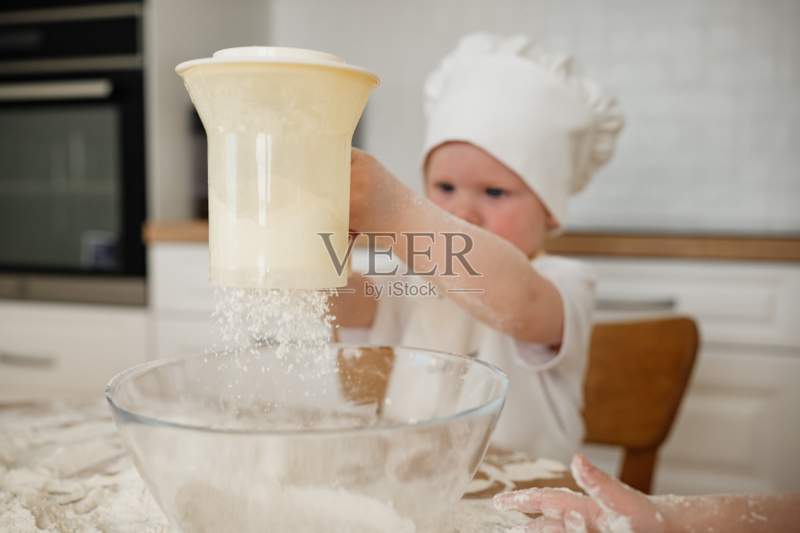 可爱的小男孩戴着厨师的帽子，围着围裙，在厨房里用筛子筛面粉照片摄影图片