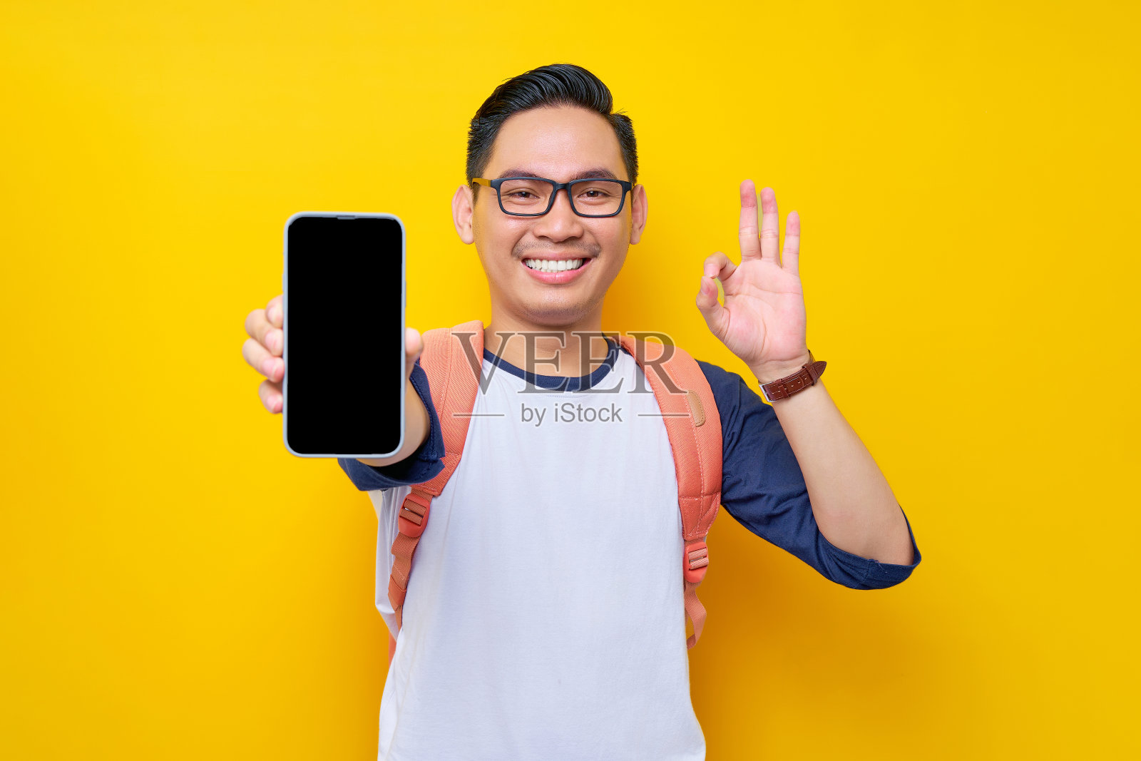 微笑着的年轻亚洲学生，穿着休闲服，戴着眼镜，背包上显示着智能手机的空白屏幕和黄色背景上孤立的ok标志。高中教育大学学院理念照片摄影图片