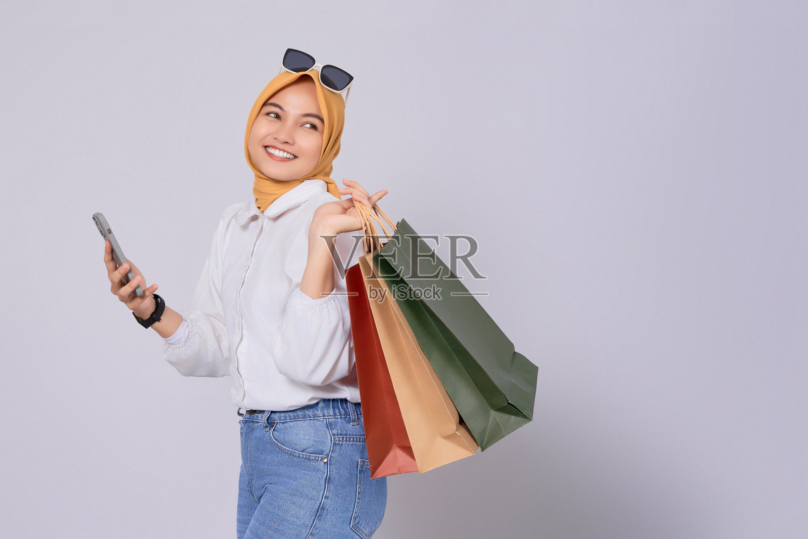 美丽的微笑年轻的亚洲穆斯林妇女拿着手机和购物袋孤立的白色背景。网上移动购物概念照片摄影图片