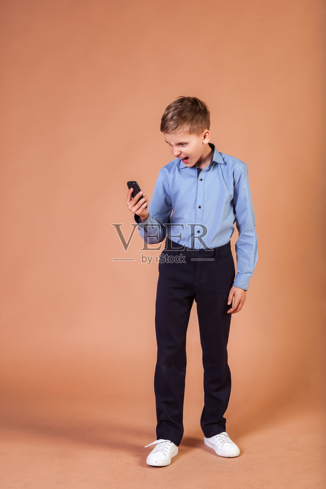 愤怒的10岁男生，穿着蓝色衬衫，拿着米色背景的手机，看着手机。男模特用手机，嘴巴尖叫。孩子情感，教育观念。复制广告文本空间照片摄影图片