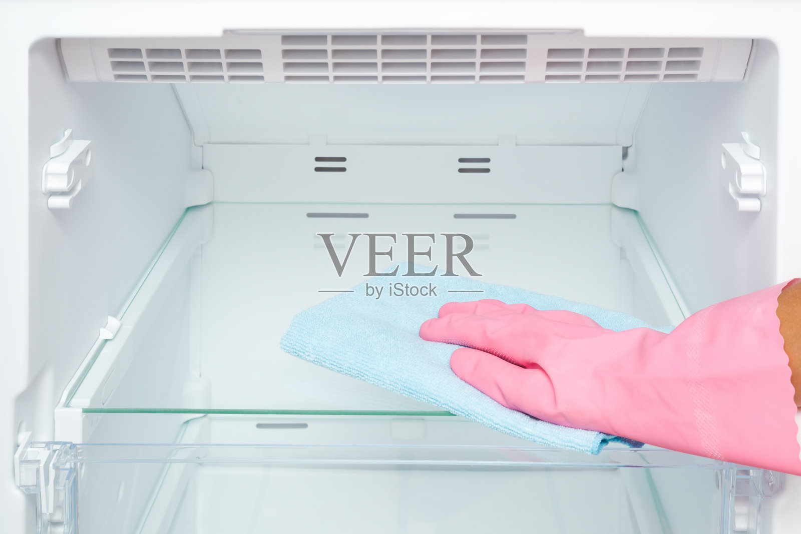 女士手戴粉色橡胶防护手套，用蓝色干超细纤维抹布擦拭冰柜的玻璃架子。特写镜头。视角镜头。照片摄影图片