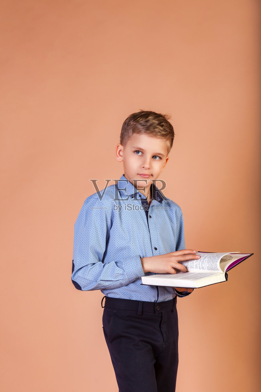 米色背景下，男生模特手里拿着书，若有所思地望向别处。10岁男生的肖像，穿着蓝色衬衫，拿着书。孩子学习，教育理念。复制广告文本空间照片摄影图片