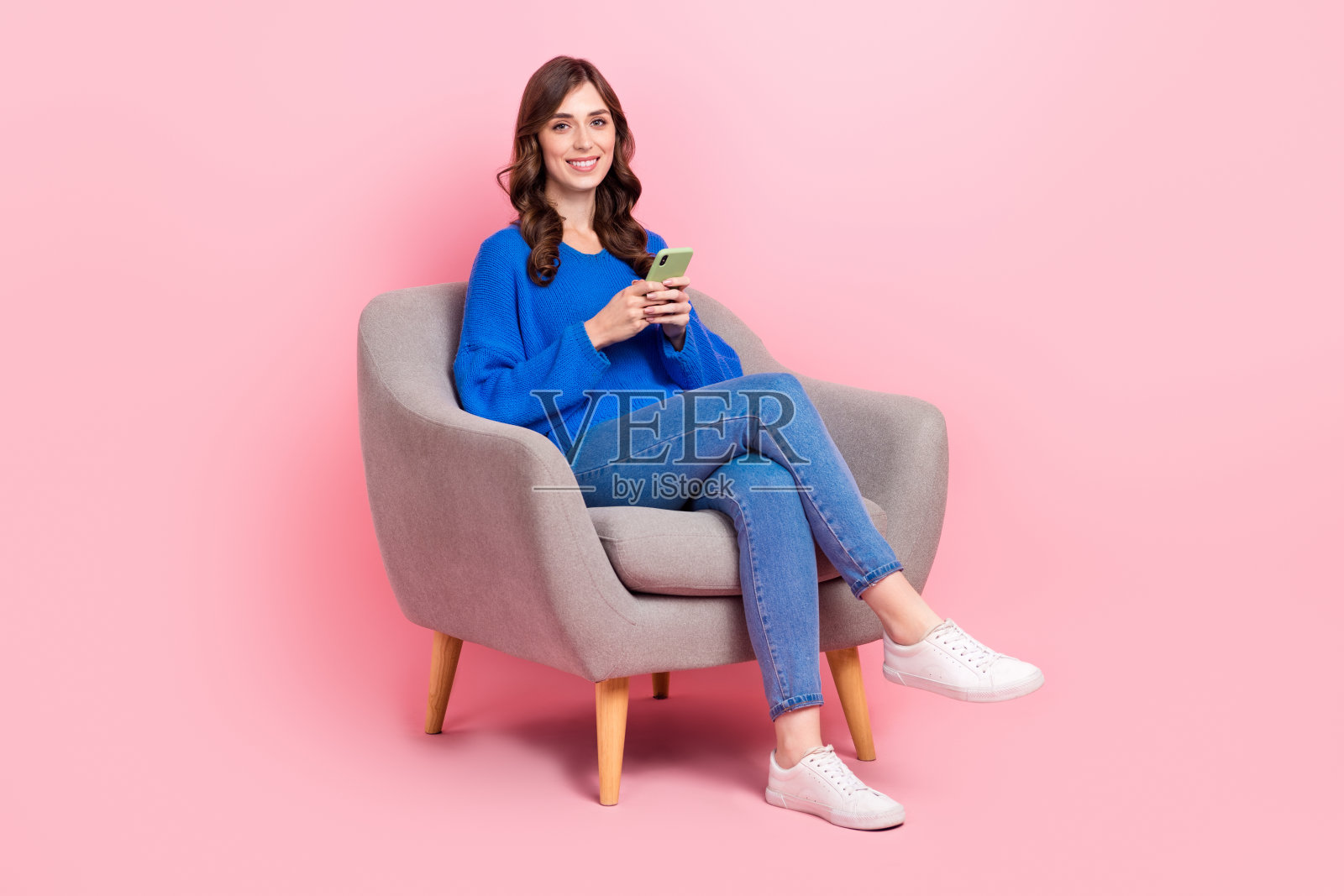 漂亮年轻女孩的全身照片，坐在扶手椅上，拿着小工具，放松地穿着时髦的蓝色衣服，孤立在粉红色的背景上照片摄影图片