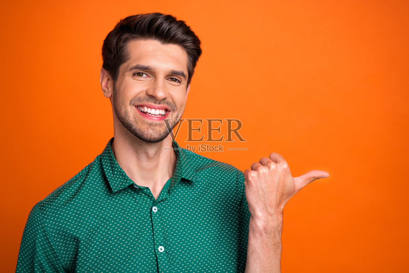 照片中开朗可爱的小伙子穿着绿色的衬衫，用大拇指指着空旷的空间，孤立的橘黄色背景照片摄影图片