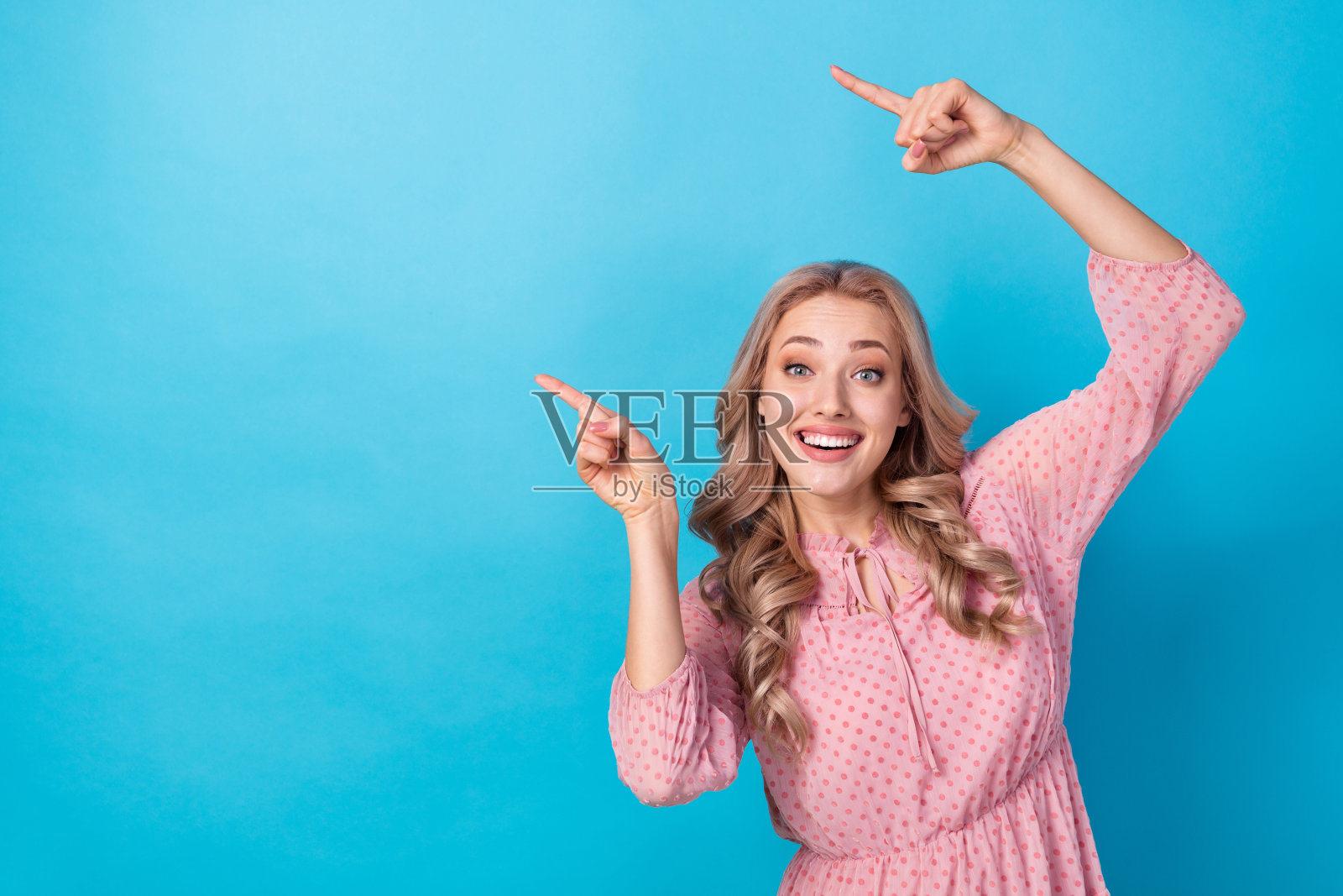 印象深刻的卷发女孩的肖像，穿着粉红色的衬衫，指向命题的空白空间，孤立在蓝色背景上照片摄影图片