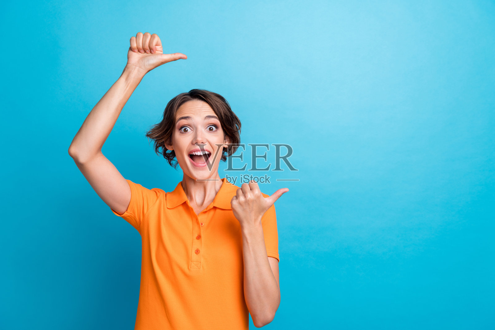 照片中开朗兴奋的女孩穿着橙色马球，张开嘴巴，露出两个大拇指，空白的空间孤立的蓝色背景照片摄影图片