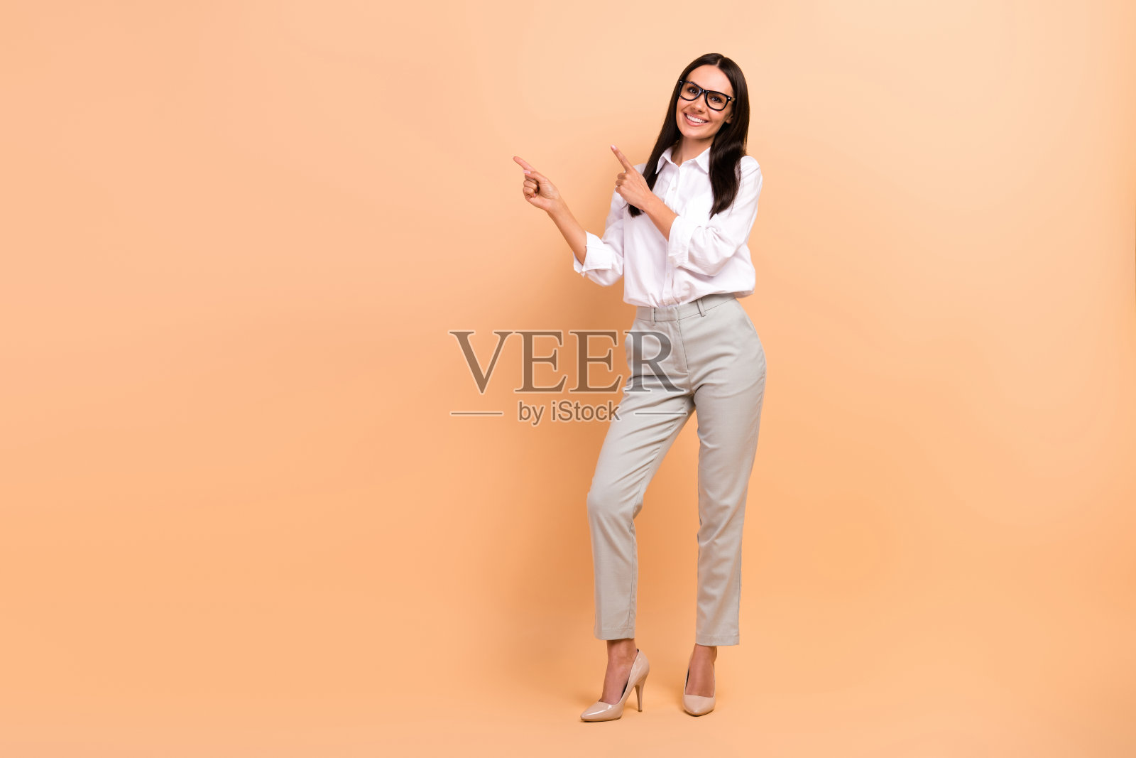 全身照片快乐微笑的女人穿着白衬衫，戴着眼镜指着两个手指空白空间孤立米色背景照片摄影图片