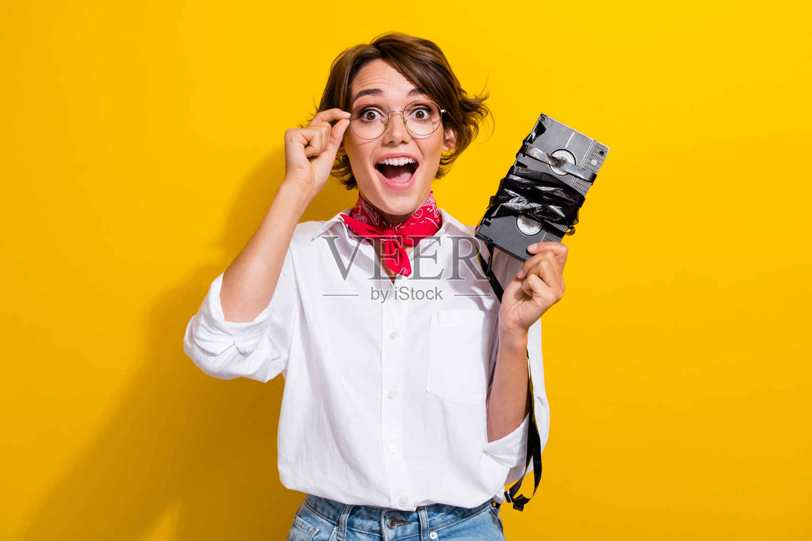 印象深刻的漂亮女士的肖像，手臂触摸眼镜，张开嘴，持有磁带录像带孤立的黄色背景照片摄影图片