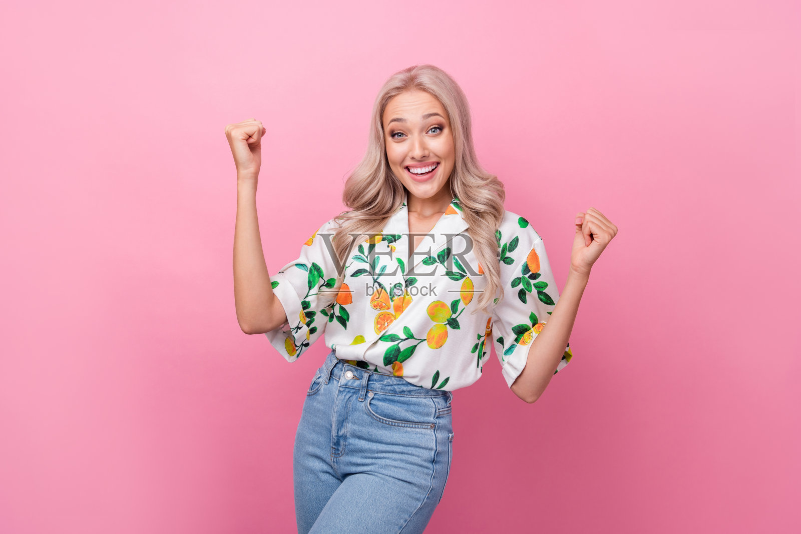 肖像的喜出望外的少女与波浪的头发，穿着时髦的衬衫紧握拳头赢得赌注孤立的粉红色背景照片摄影图片