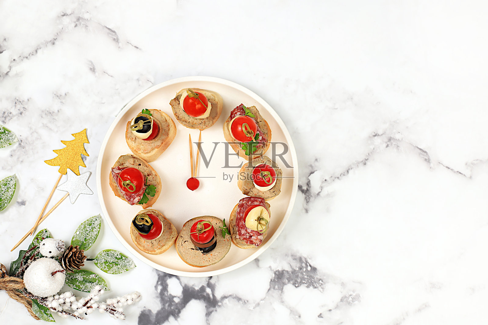 圣诞和新年的菜肴，一套小吃的节日餐桌。时钟形状的盘子canapÃ©由西红柿、肉酱、马苏里拉奶酪、意大利腊肠、橄榄、黑橄榄和葡萄在大理石桌子上制成，照片摄影图片