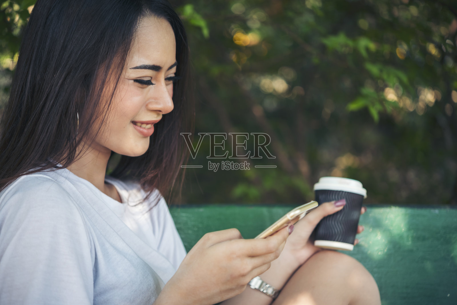 一名亚洲女子在绿色公园用智能手机喝咖啡，手拿热一次性杯子。横幅快乐放松亚洲妇女笑脸在户外花园。年轻女性享受大自然的复印空间照片摄影图片