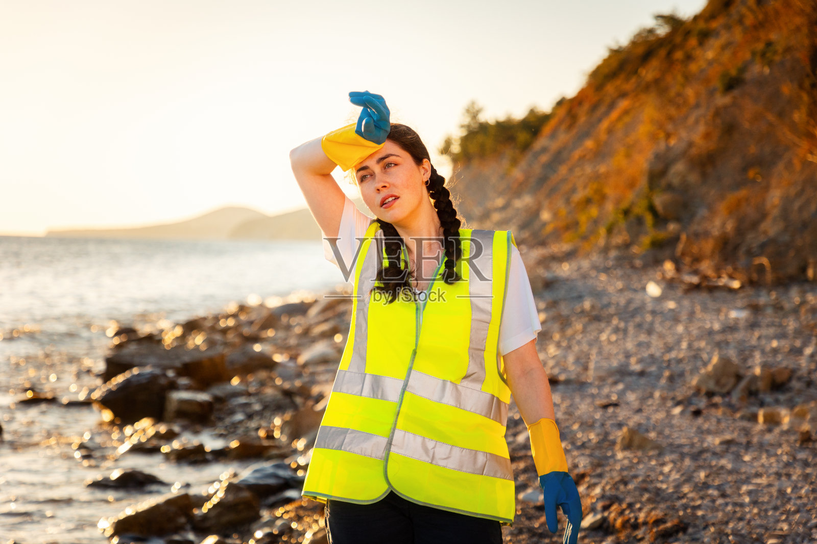 疲惫的年轻志愿者穿着背心，戴着手套擦拭额头上的汗水。大扫除日。海洋污染与环境保护的概念照片摄影图片