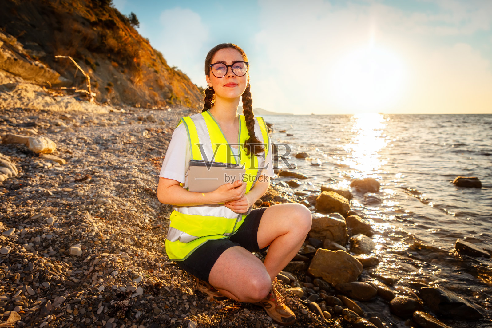 地球日。微笑的女人穿着背心，拿着电子平板电脑蹲在海边的海滩上。海洋污染与环境保护的概念照片摄影图片