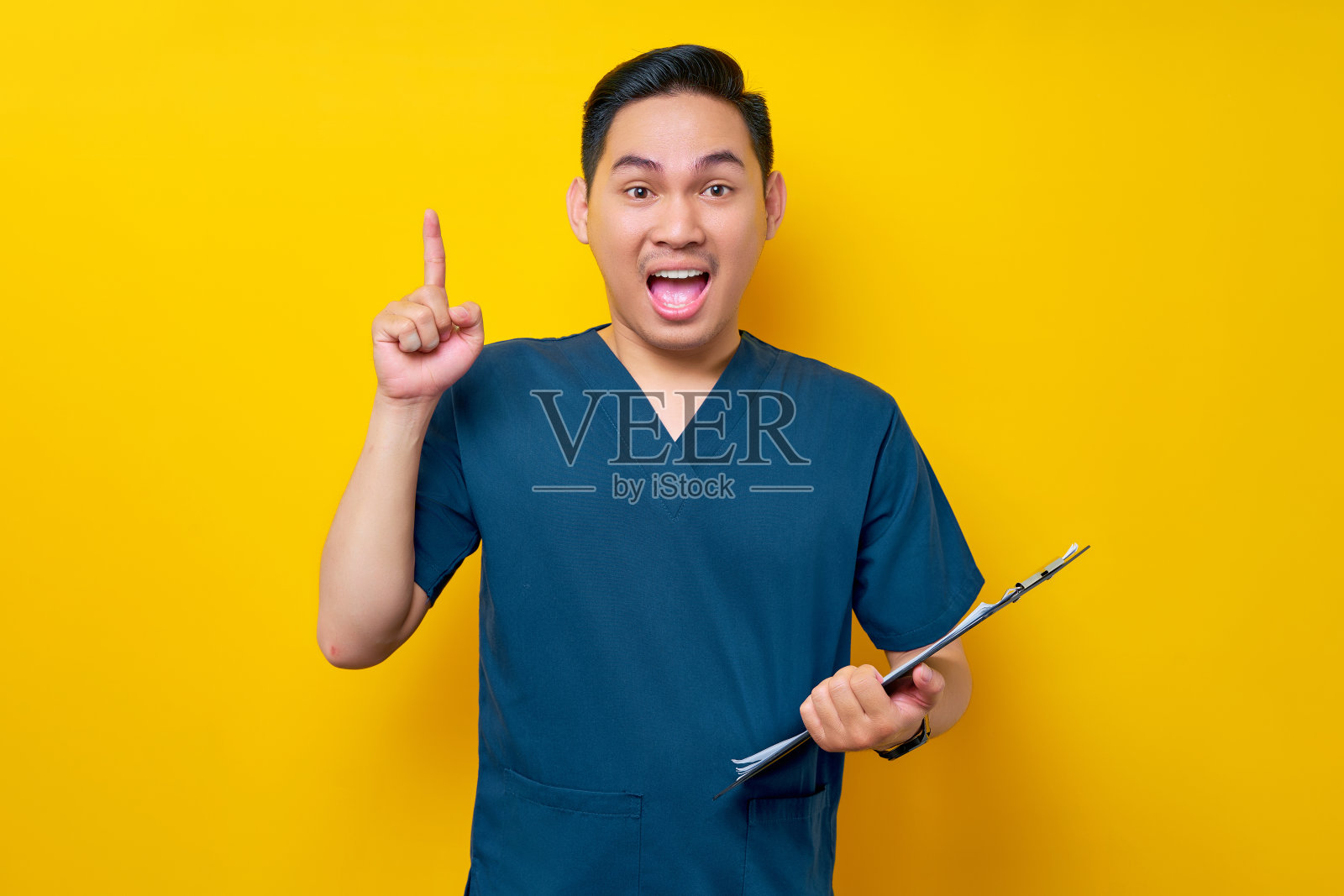 穿着蓝色制服，兴奋的年轻亚洲专业男医生或护士，拿着写字板，手指向上，在黄色的背景上有伟大的想法。保健医学概念照片摄影图片