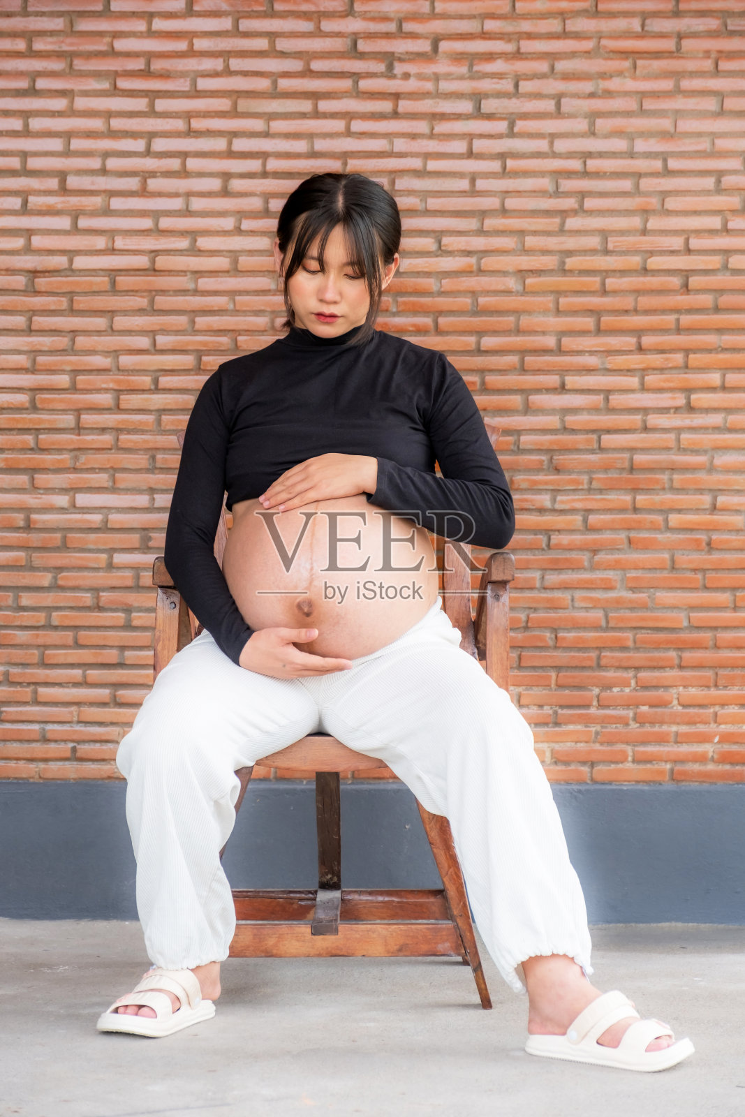 穿着裙子的孕妇妈妈在红砖背景上把手放在肚子上，用爱等待着你的宝宝的诞生照片摄影图片