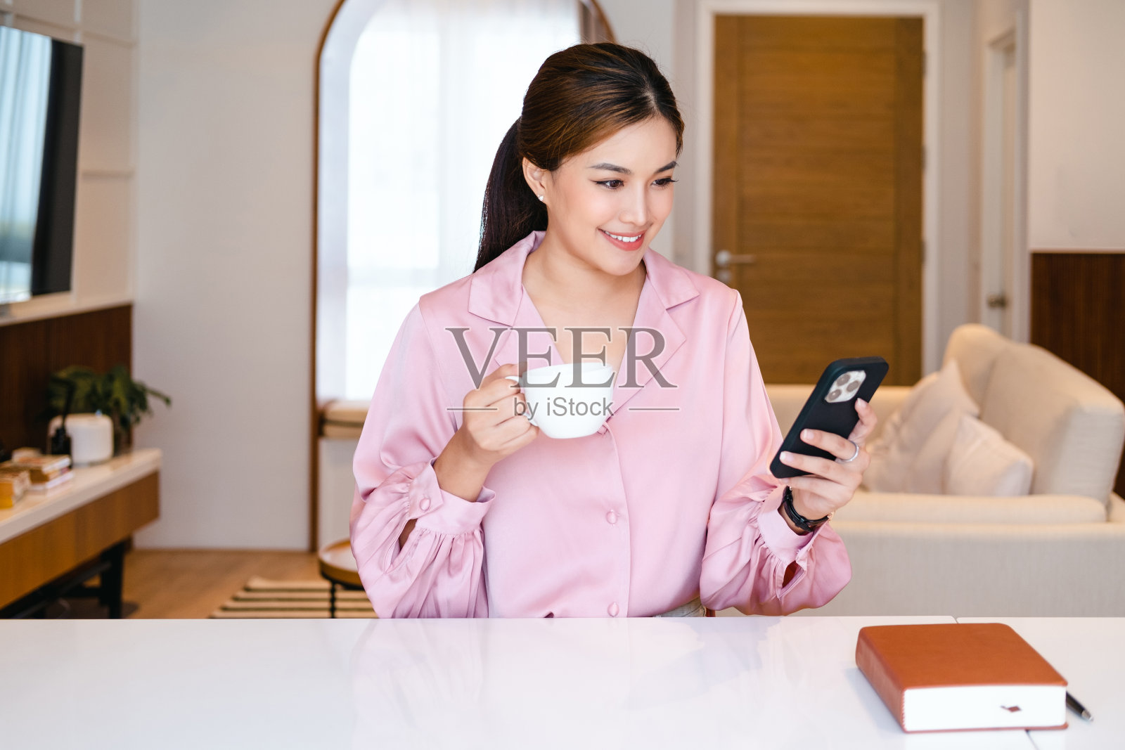 年轻漂亮的女人拿着咖啡杯，坐在家里办公室看智能手机。

快乐的女大学生用手机。女商人微笑着喝着咖啡。照片摄影图片