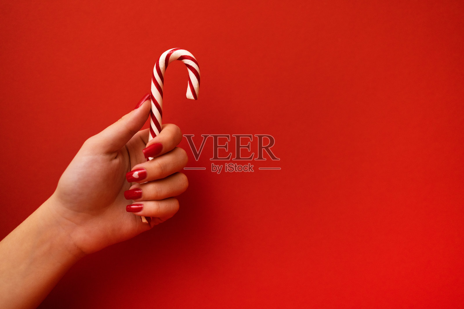 一个涂着红色指甲的年轻女孩手里拿着一根拐杖糖。肉桂或薄荷棒在明亮的红色背景。照片摄影图片