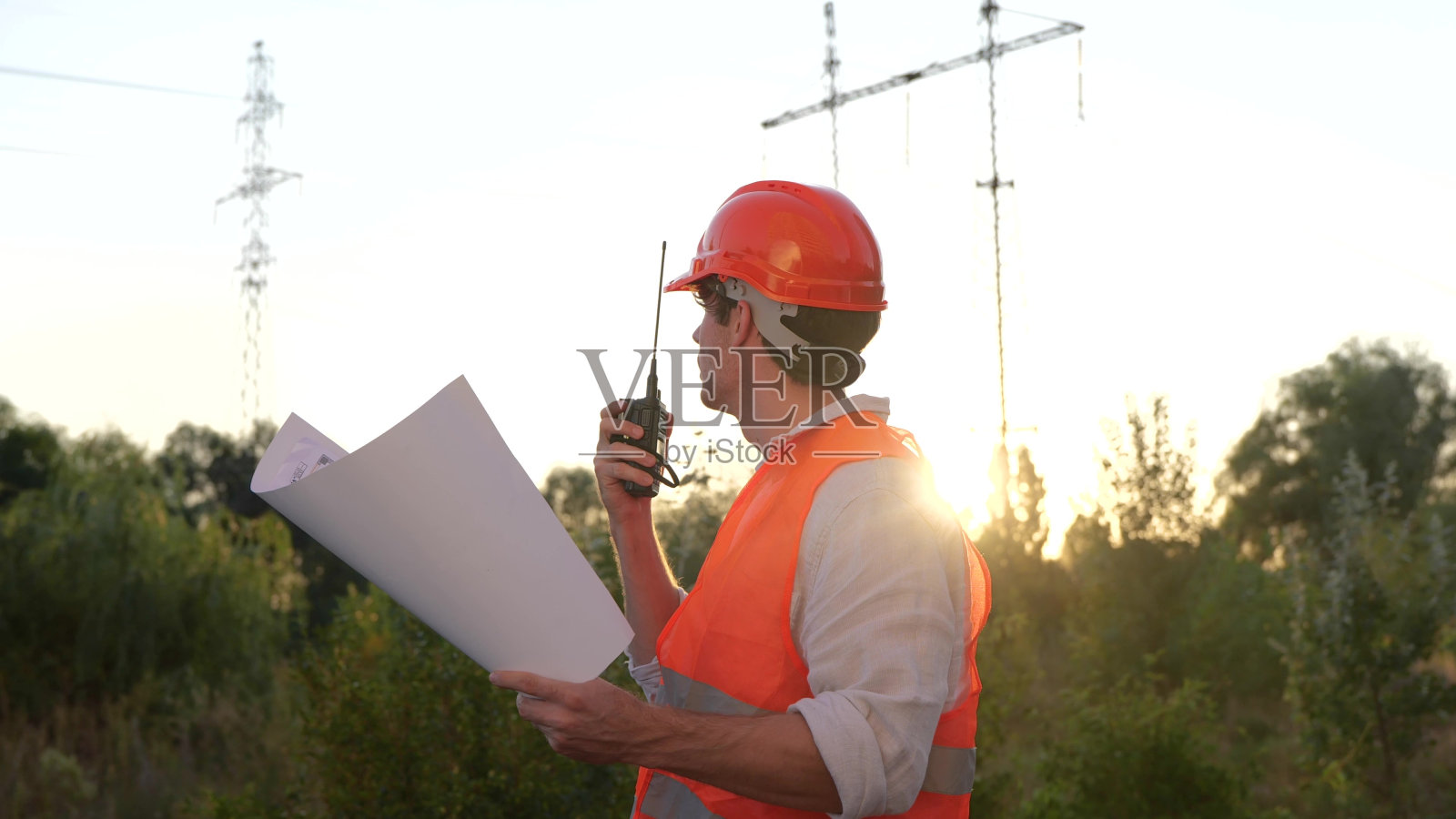 电气工程师手持图纸在对讲机上讲话，同时控制室外高压电塔的维修。能源工业概念。慢动作照片摄影图片
