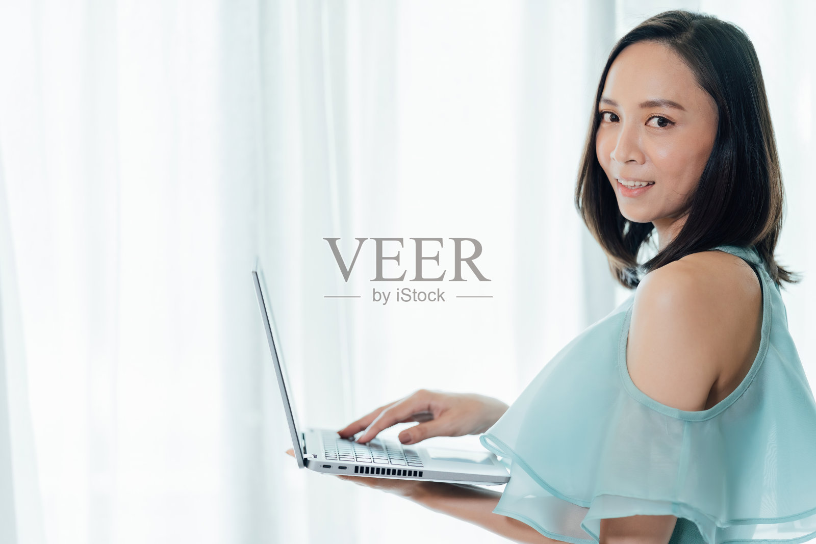 该公司的亚洲女商人领袖站在办公室里，拿着笔记本电脑，微笑着看着对方的眼睛，正在做一个大型的在线营销项目。照片摄影图片