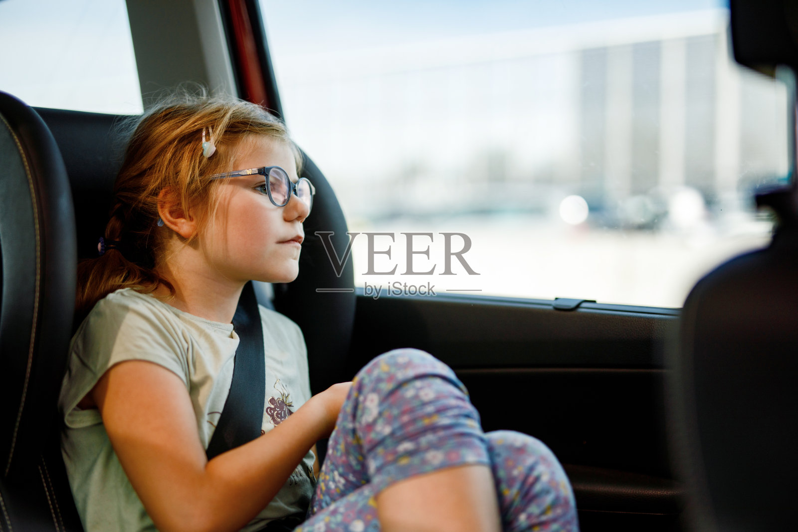 学龄前的小女孩坐在她的汽车座椅上。戴眼镜的快乐孩子望着窗外。在交通堵塞的家庭度假路上微笑的孩子。照片摄影图片