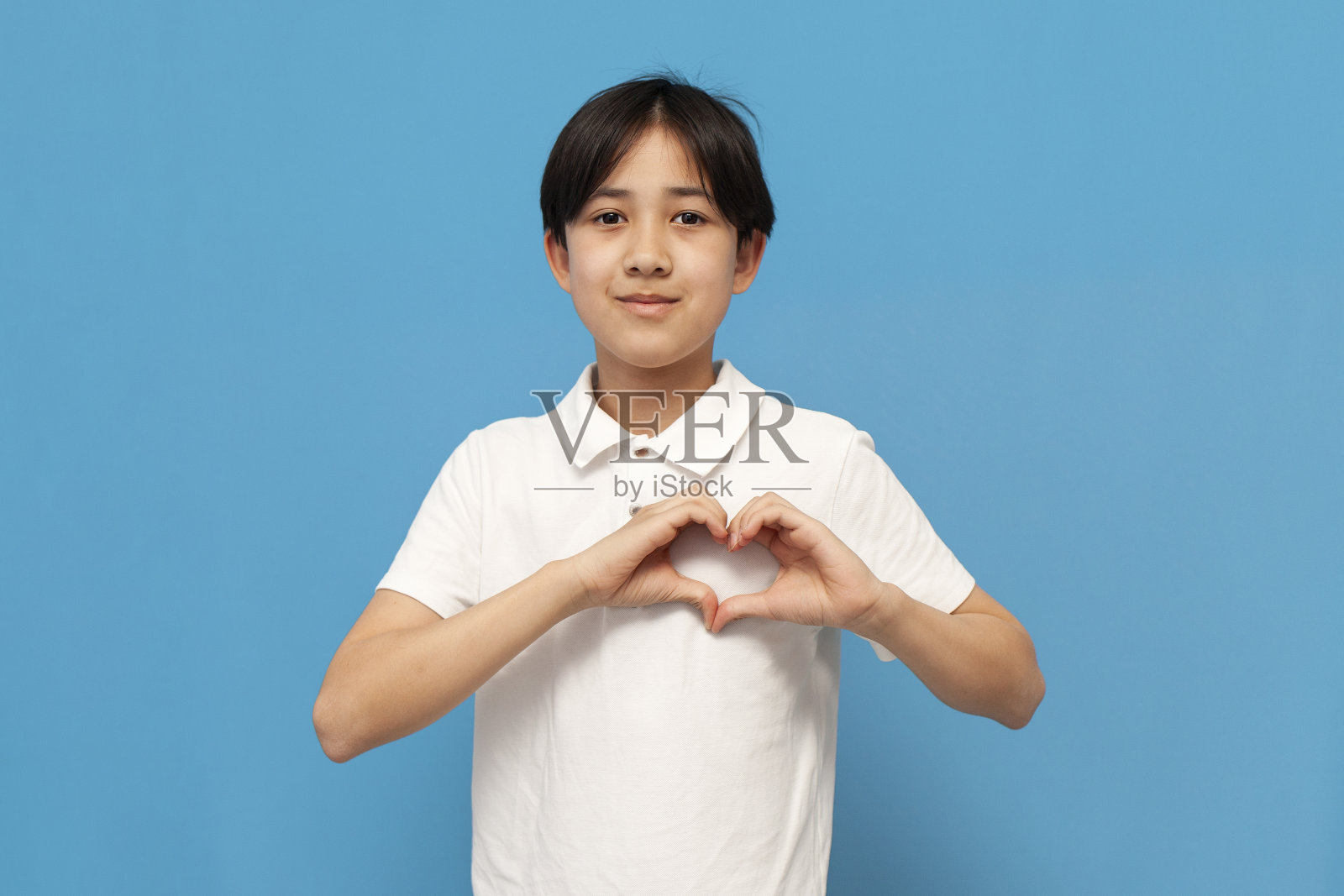 12岁的亚洲男孩穿着白色t恤，双手放在蓝色孤立的背景上，展示了他的心照片摄影图片