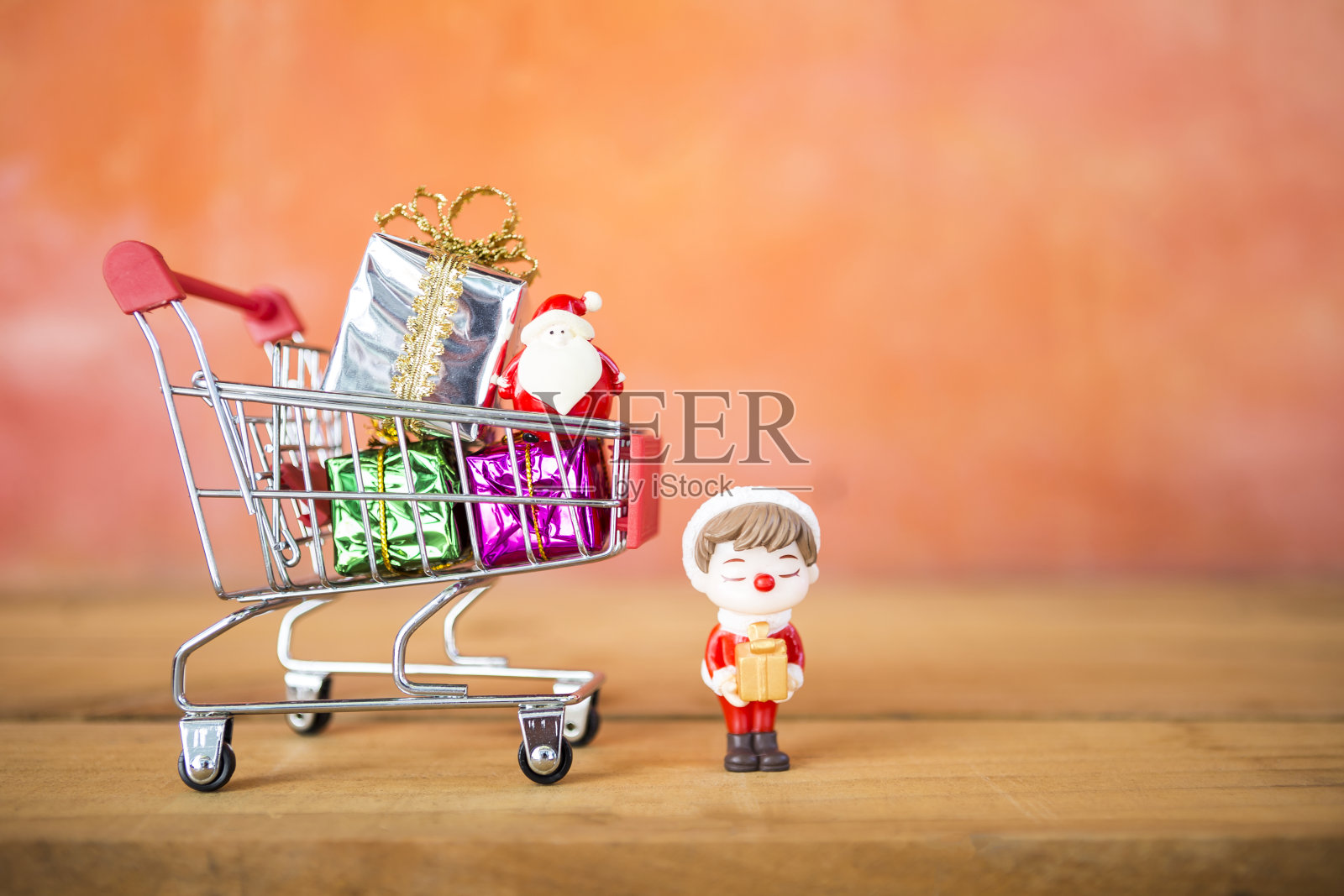 礼盒与圣诞老人在购物车和微型男孩拿着礼物与空间模糊的橙色墙壁背景照片摄影图片