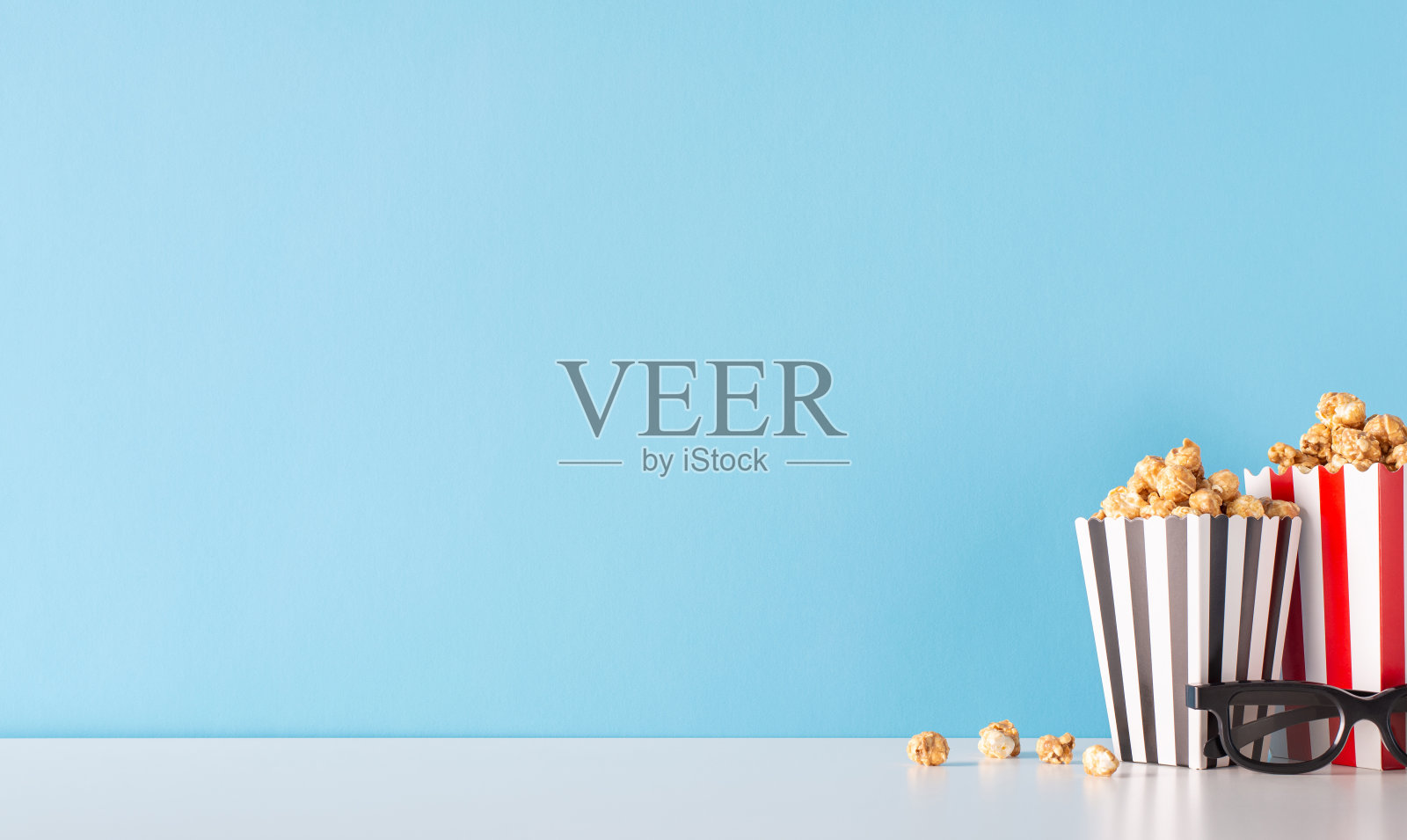 展示你的电影首映与美味的治疗主题。侧视图的桌子装饰着3D眼镜，和美味的爆米花在条纹容器对着柔软的蓝色墙壁，完美的电影宣传照片摄影图片