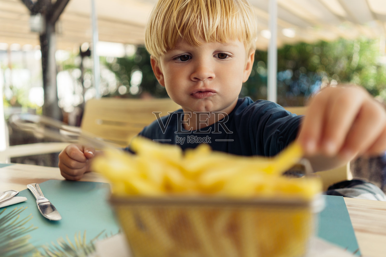 一个三岁的金发小男孩正在咖啡馆里吃薯条。照片摄影图片