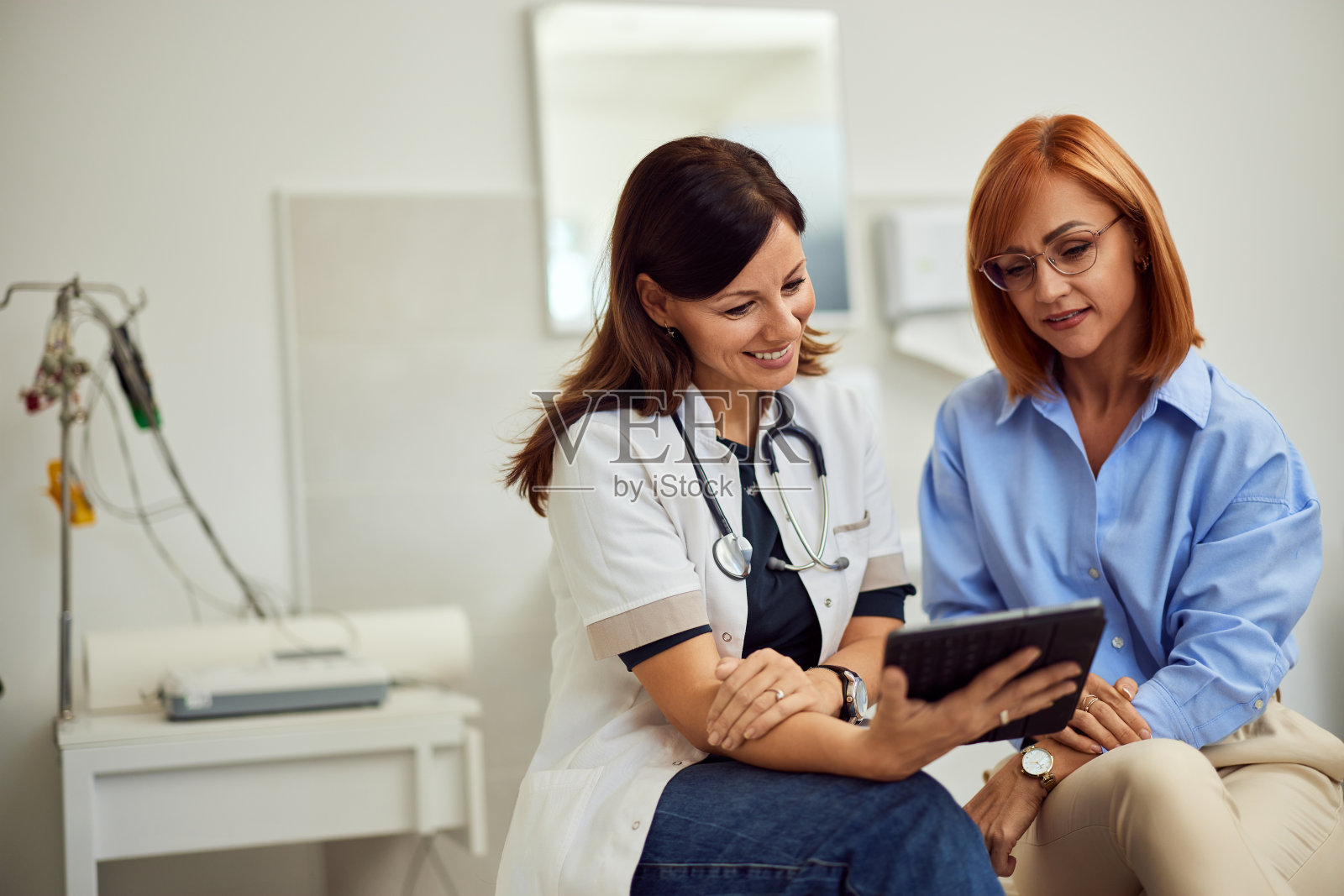 一个面带微笑的黑发医生，坐在床上和一个姜黄色头发的病人谈论她的结果。照片摄影图片