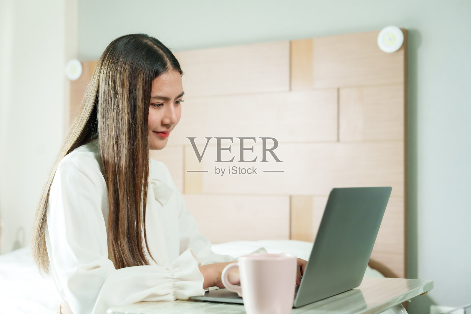 当在家工作时，美丽的长发亚洲女人坐在笔记本电脑上思考和输入电子邮件。在床上在卧室准备节假日给主管发会议纪要。照片摄影图片