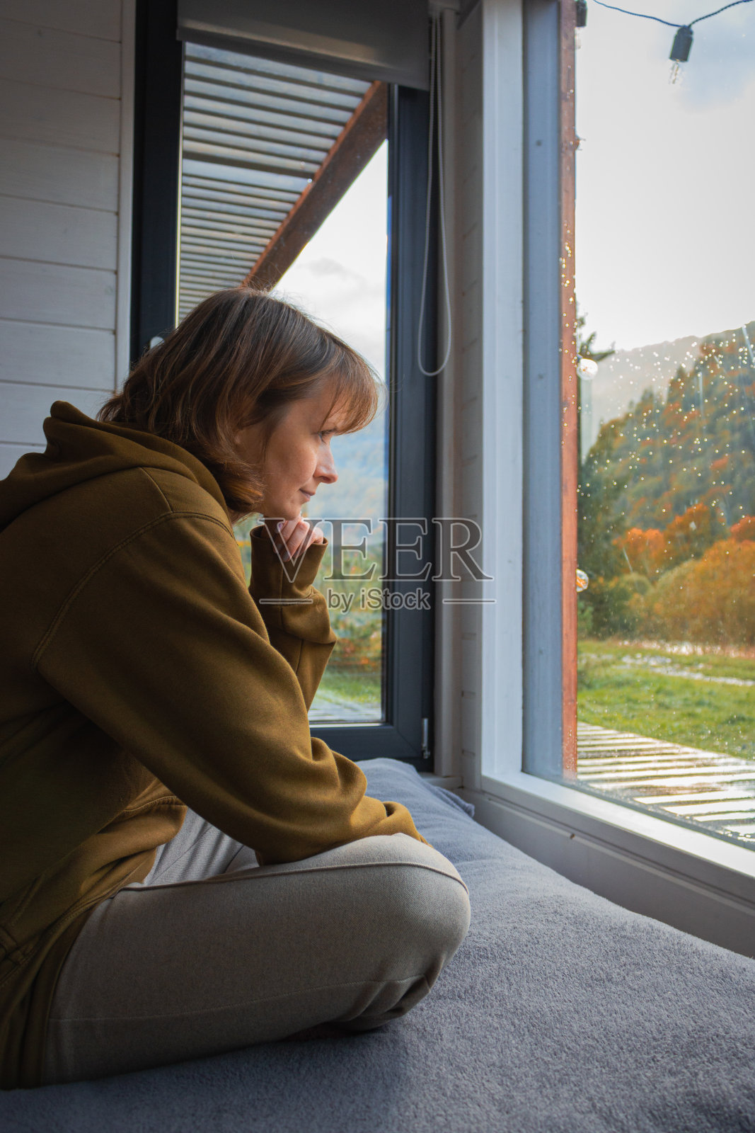 女孩坐在大窗户后面的床上，可以看到秋天的山景。窗台上穿连帽衫的女人。逃到山间小屋。在乡间小屋休闲。秋天旅游。照片摄影图片