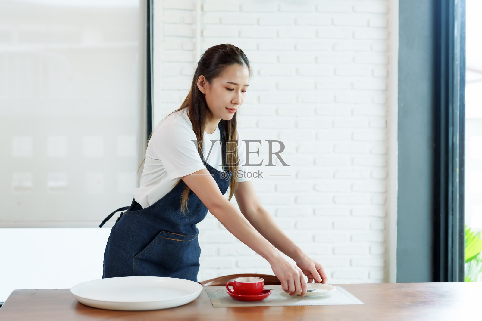 咖啡厅亚洲女员工的手正在把餐桌上用过的盘子和玻璃杯收集到托盘里。在小咖啡店要把桌子打扫干净，为下一批顾客使用服务做准备。照片摄影图片