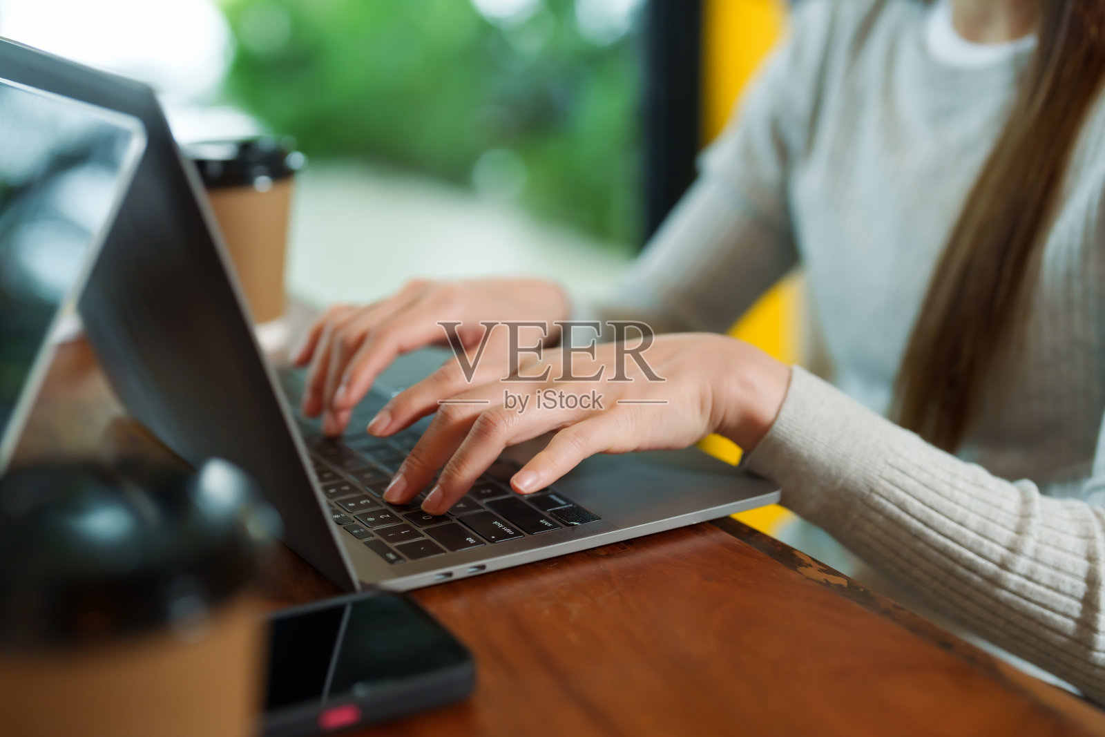 亚洲女商人的手在小咖啡馆打字和检查销售情况，木制桌子上放着笔记本电脑，旁边是棕色的一次性纸质咖啡杯。上面有电话。照片摄影图片