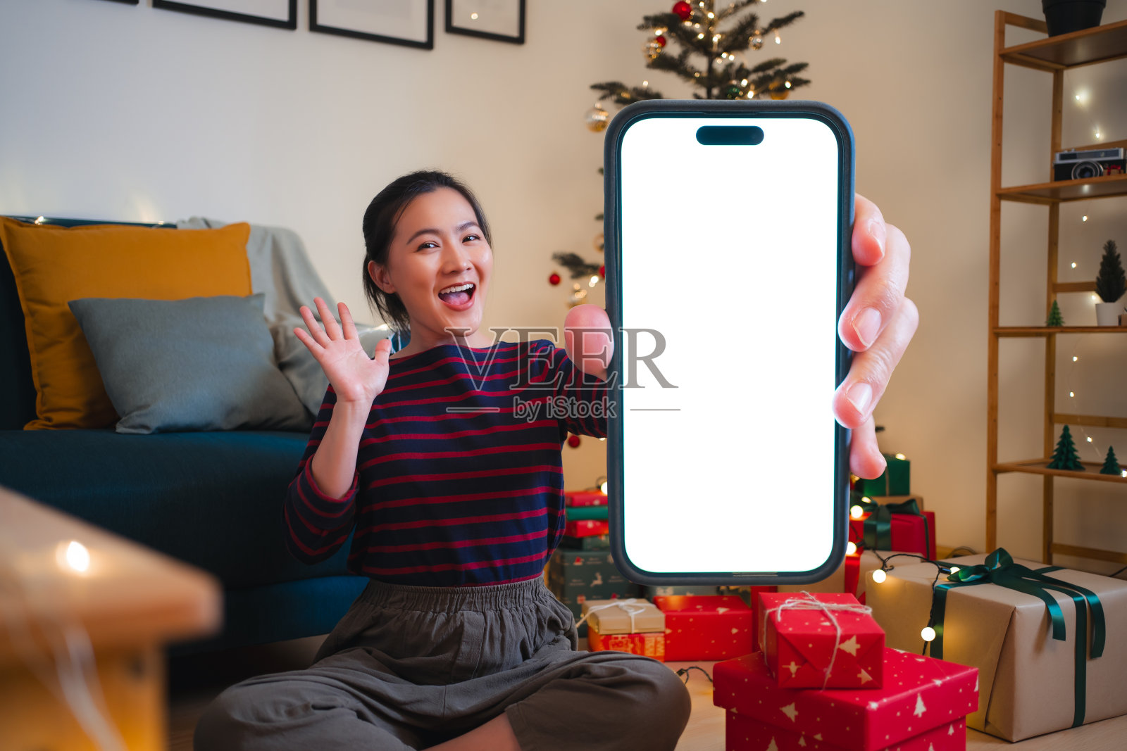 一名亚洲女性坐在装饰着圣诞树的客厅里，拿着白色屏幕的智能手机，开心而兴奋照片摄影图片
