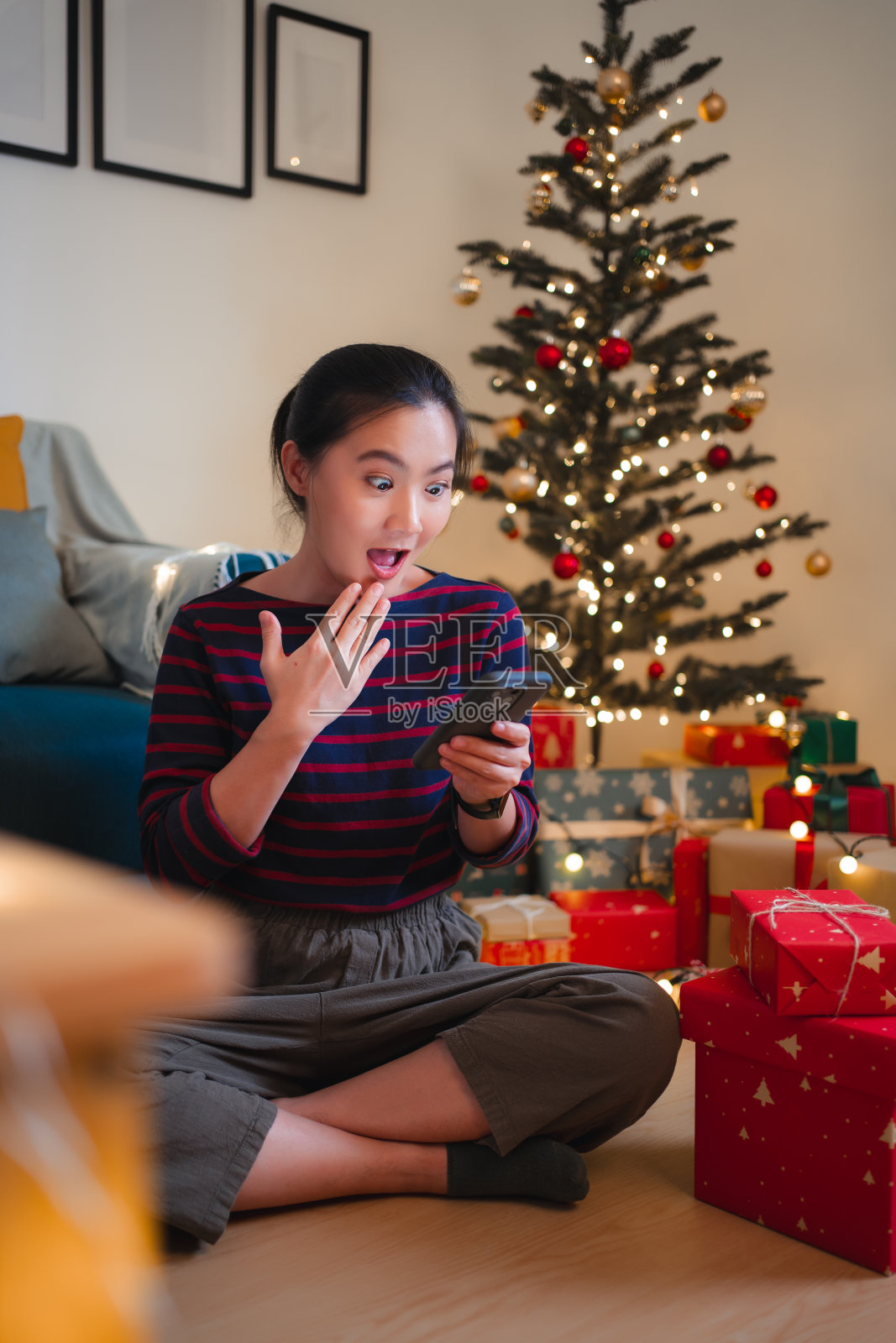 亚洲女人坐在装饰着圣诞树的客厅里，看着智能手机，感到兴奋。照片摄影图片