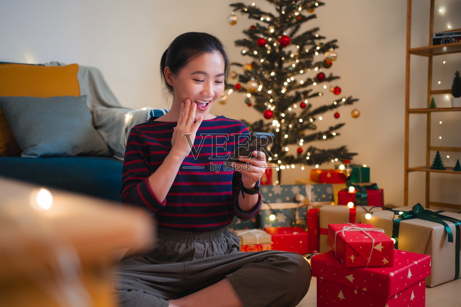亚洲女人坐在装饰着圣诞树的客厅里，看着智能手机，感到兴奋。照片摄影图片